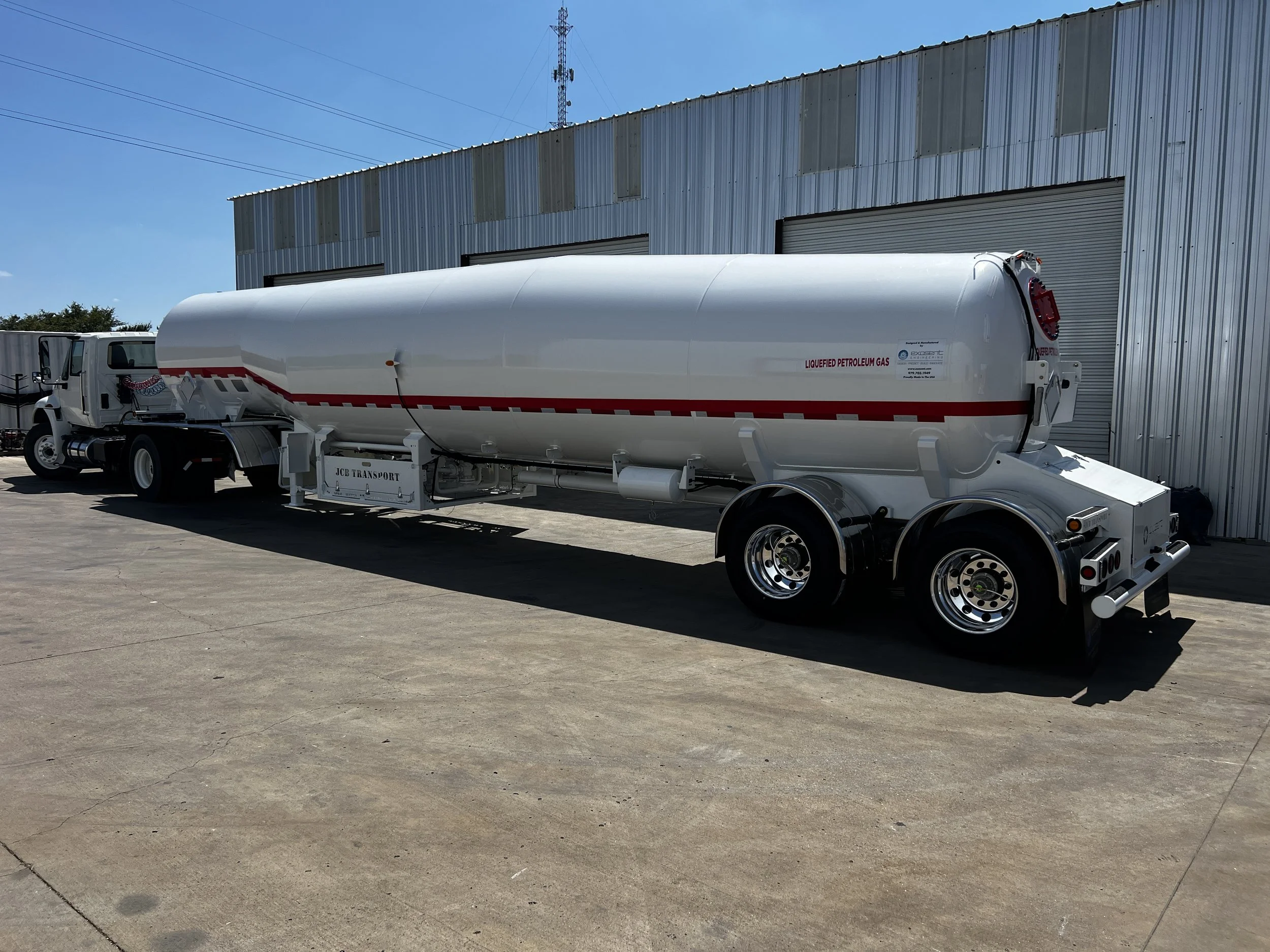White tanker truck labeled 'Liquefied Petroleum Gas' parked outside near a metal building and clear blue sky.