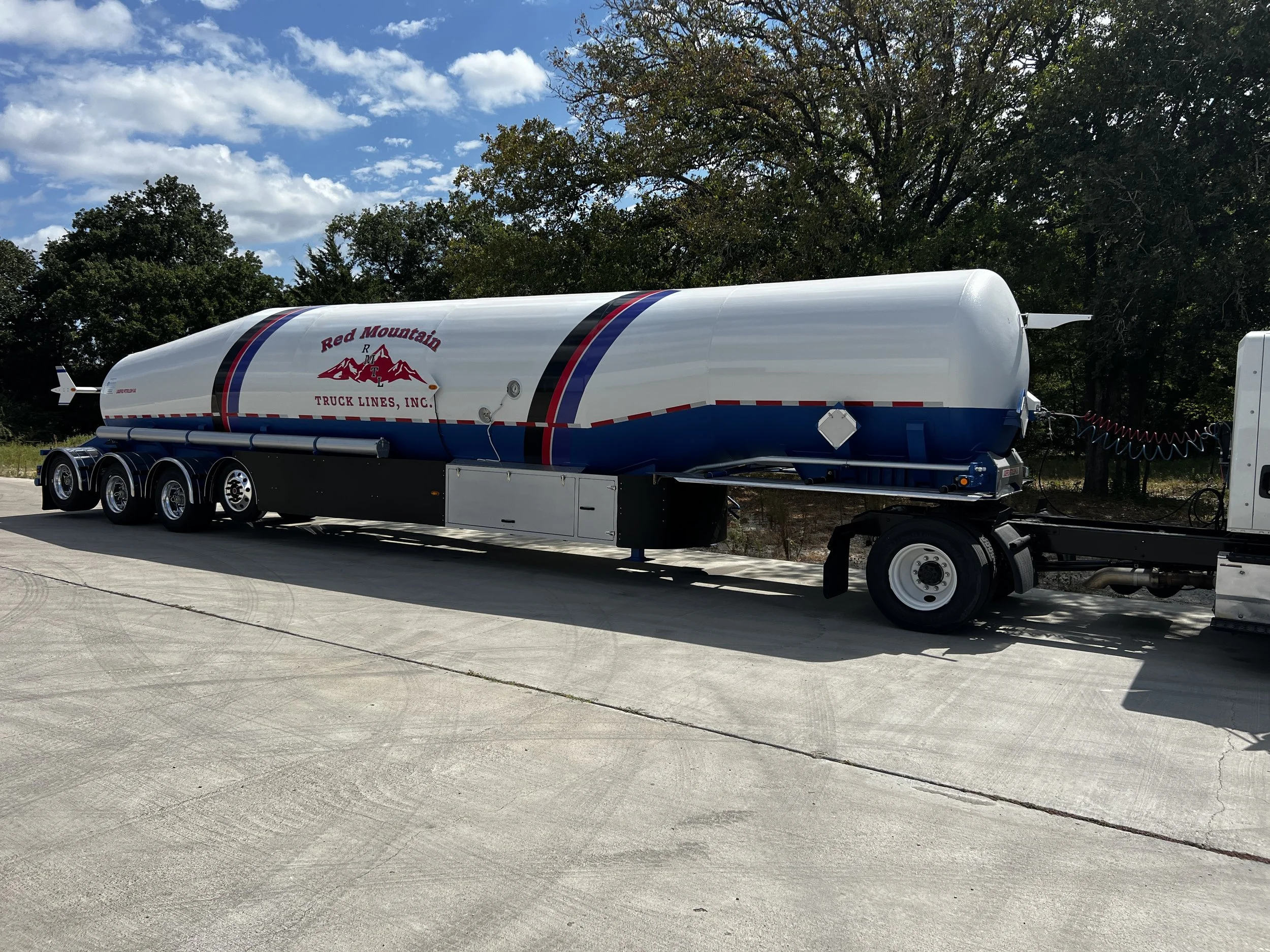 A large white and blue tanker trailer with a red mountain logo and the text "Red Mountain Truck Lines, Inc." parked on a concrete surface under a partly cloudy sky, with trees in the background.