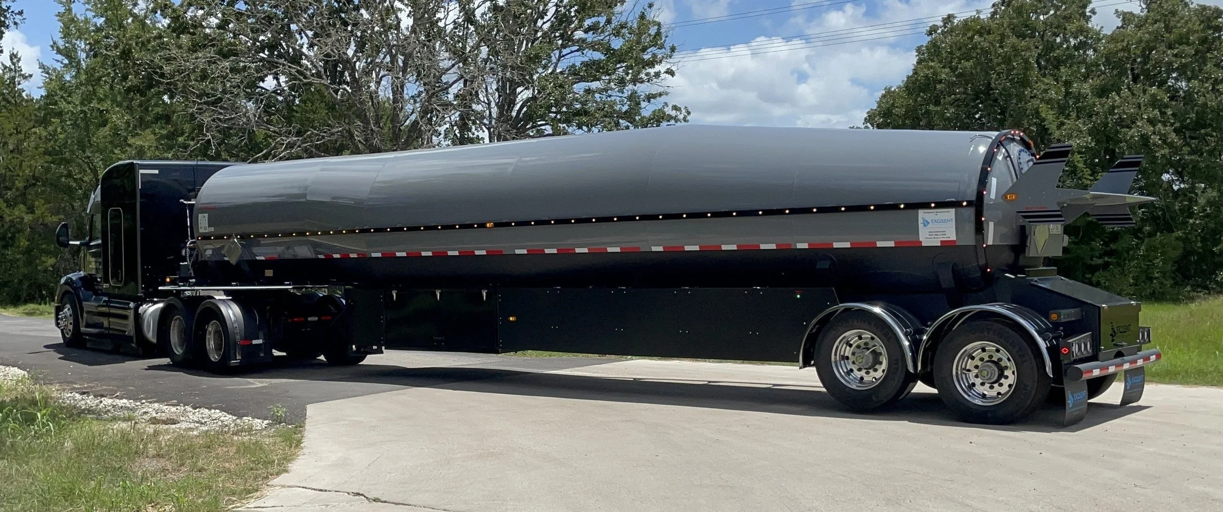 A large black tanker truck parked on a rural road, surrounded by green trees and grass, with a partly cloudy sky.