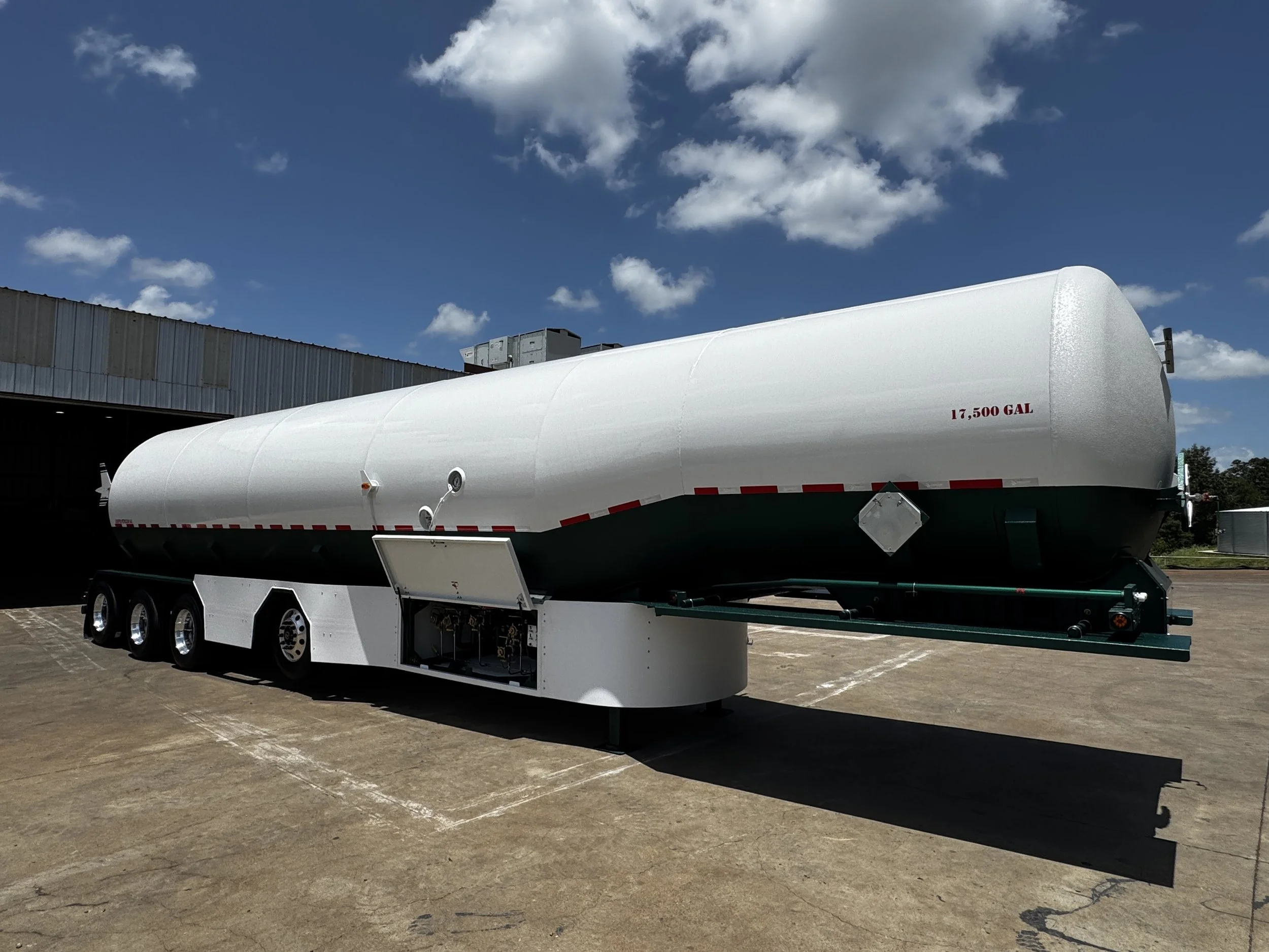 A large white tanker trailer with a capacity of 17,500 gallons is parked outdoors on a paved surface, with a warehouse-like building and a blue sky with scattered clouds in the background.