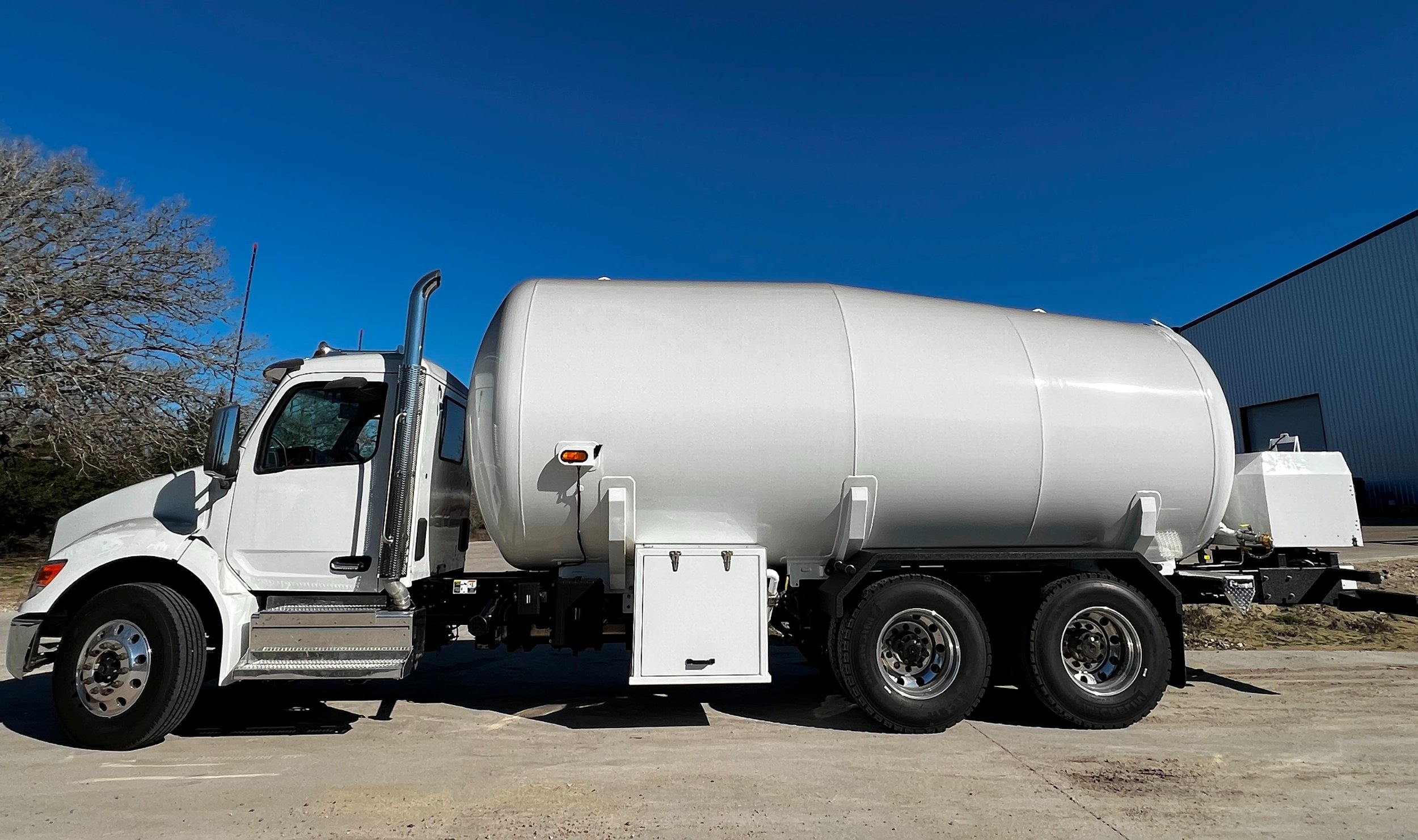 White tanker truck parked outside near a large industrial building with a bright blue sky in the background.