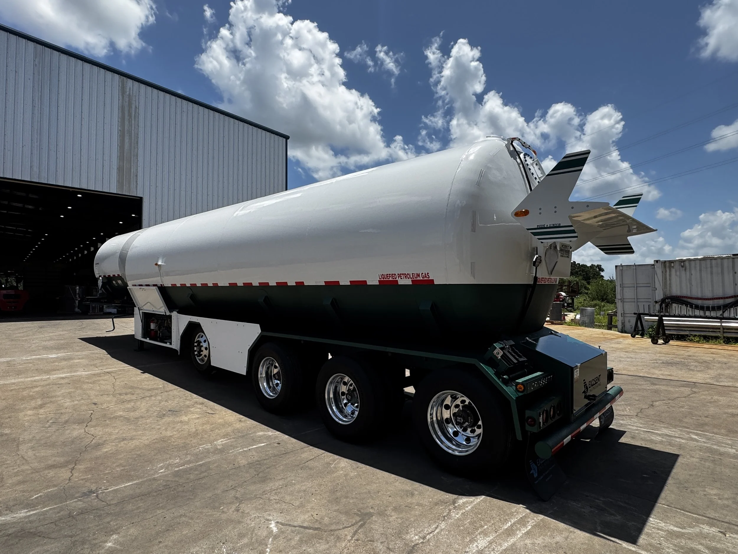 A white liquid petroleum gas storage tank mounted on a trailer parked outdoors near a large metal building with a cloudy blue sky overhead.