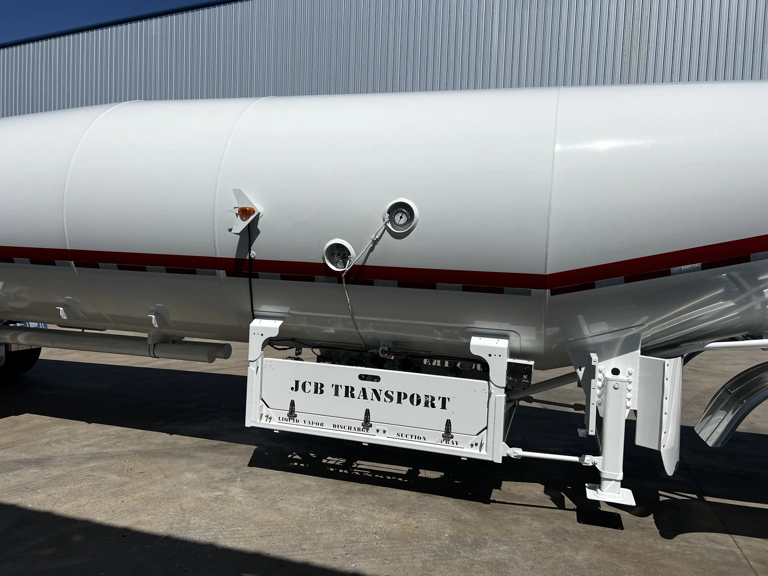 A large white tanker trailer with a red stripe, labeled 'JCB TRANSPORT' underneath, parked on a concrete surface.