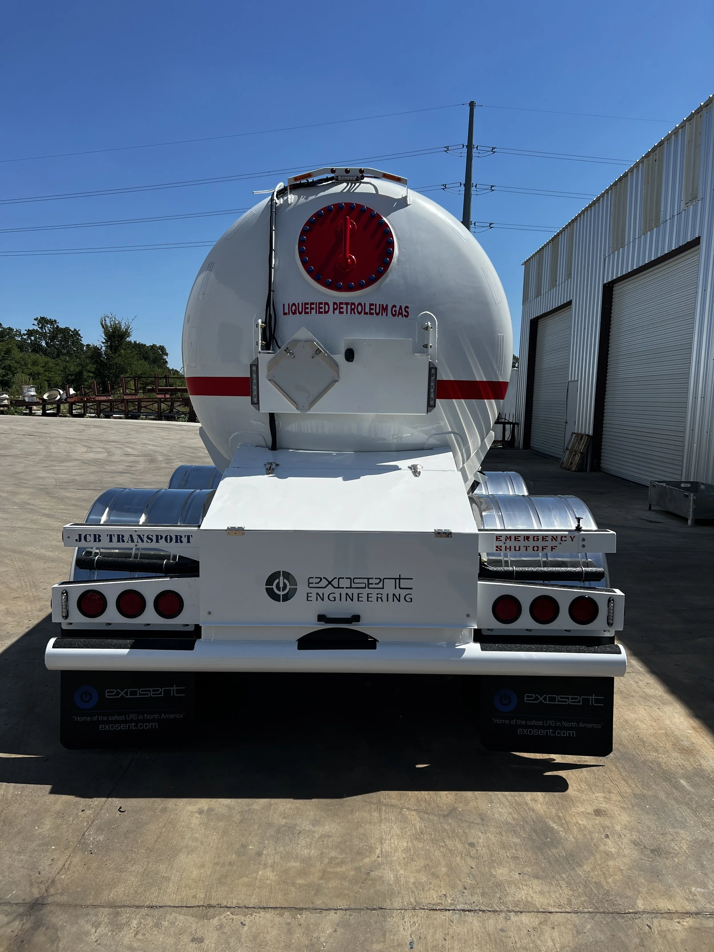 Rear view of a white liquefied petroleum gas tanker truck parked outside a warehouse with closed roll-up doors under a clear blue sky.