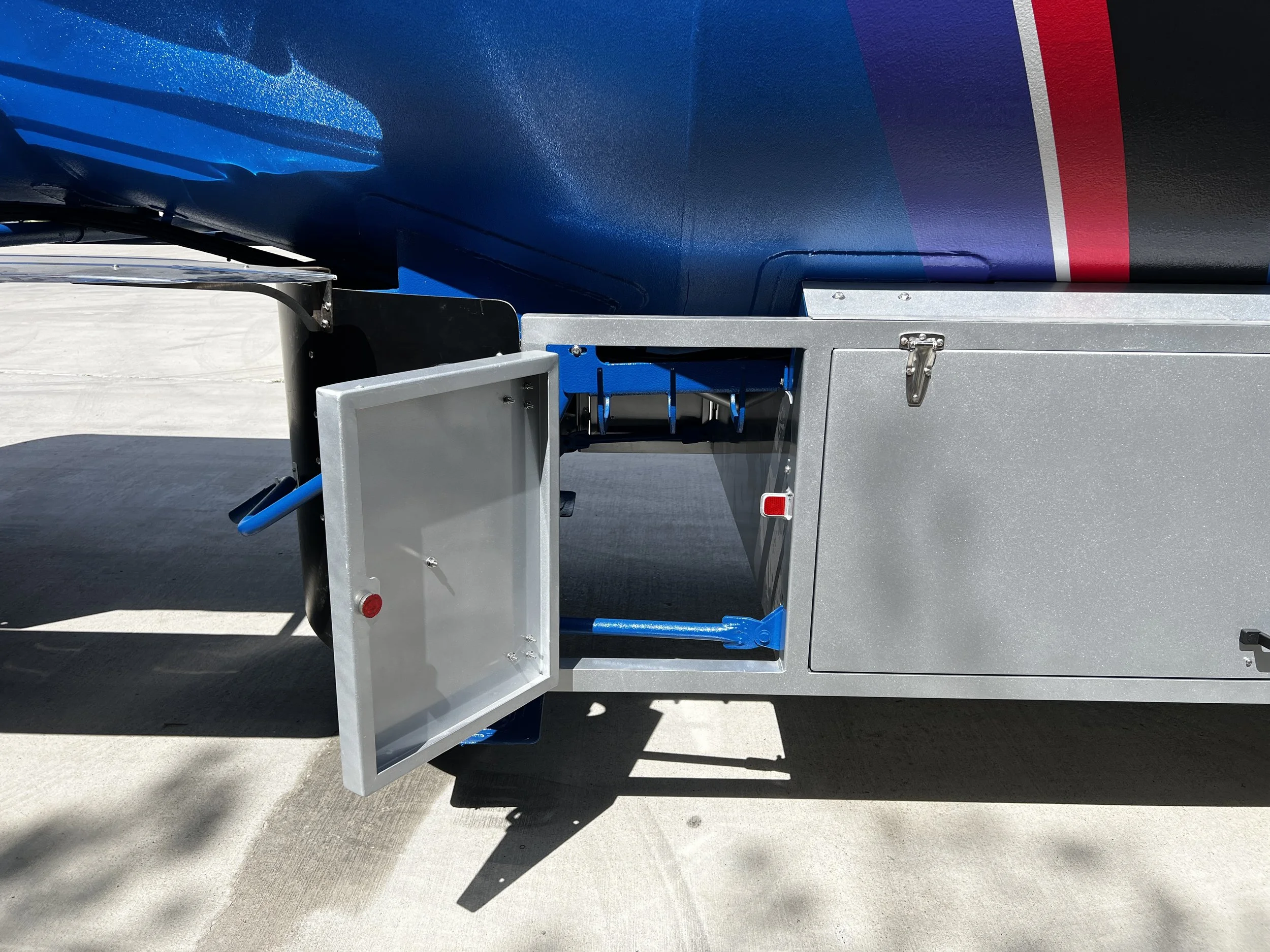 Close-up of a metallic storage compartment on an aircraft with a hinged door open, showing latches and hydraulic mechanisms, part of the fuselage with a dark blue, red, and grey painted design.