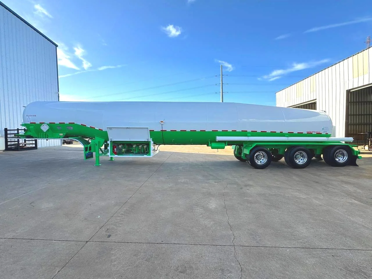 Large MC331 tanker trailer with white and green coloring parked on concrete outside industrial buildings under blue sky.