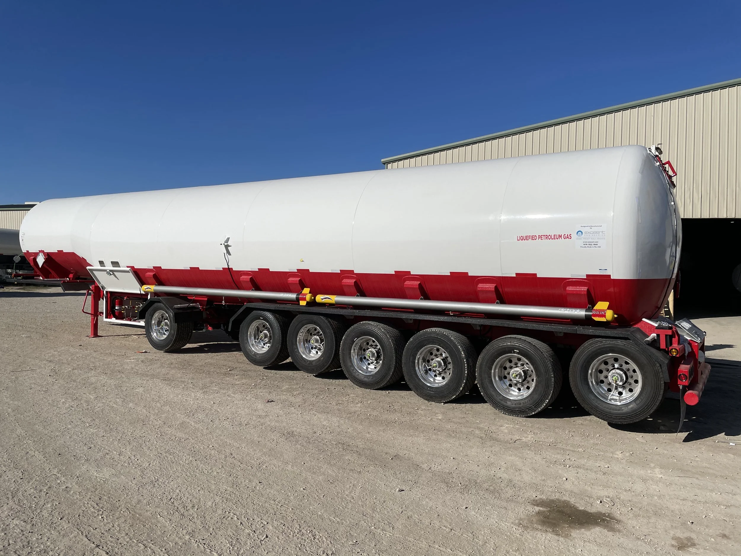 A white and red fuel tanker trailer parked outdoors on a dirt lot, with a beige industrial building in the background and a clear blue sky overhead.