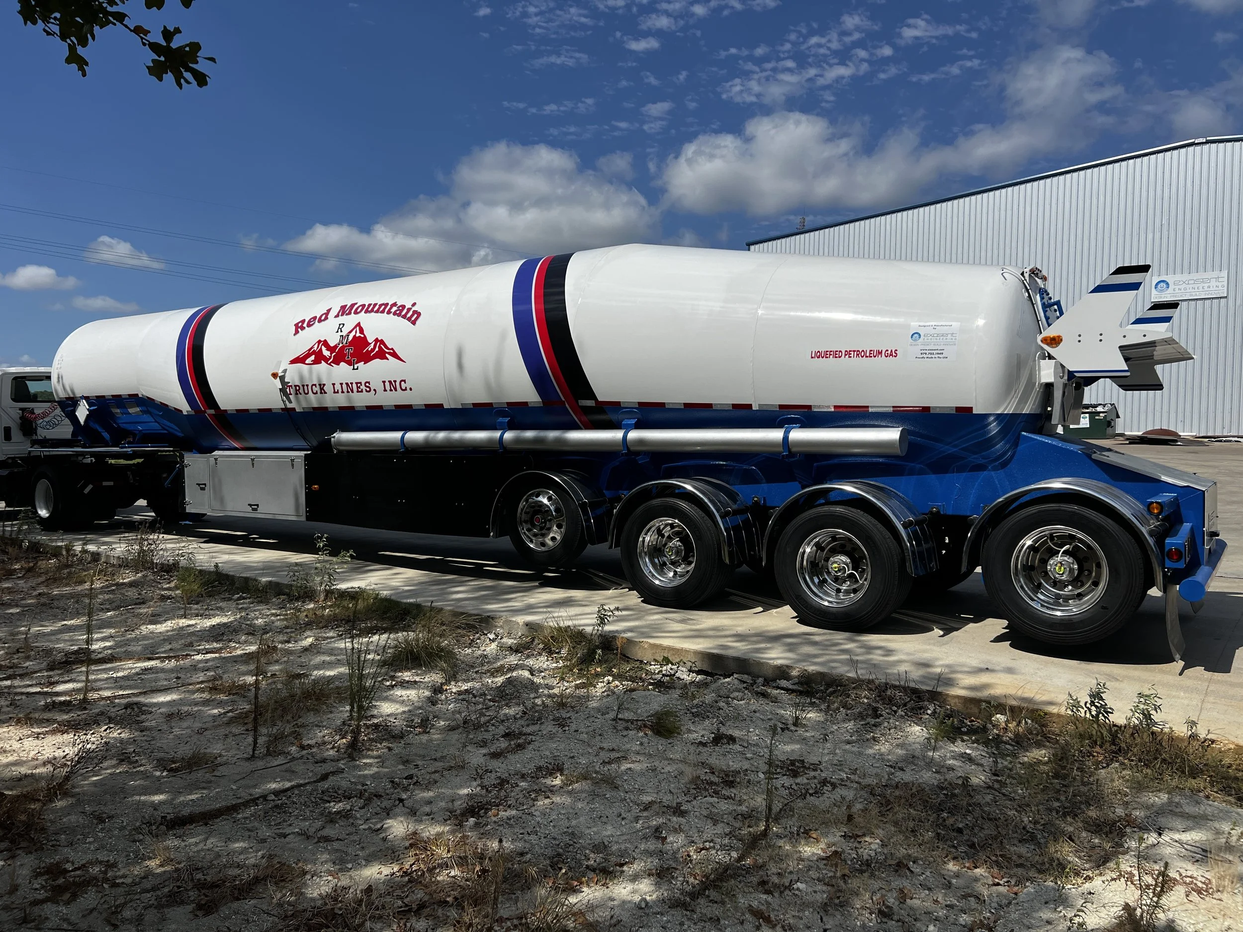 A large tanker truck parked outdoors under a partly cloudy sky. The tanker is white with red and blue stripes and has a logo that reads 'Red Mountain Truck Lines, Inc.' and a depiction of mountains. The truck is attached to a blue chassis with multip