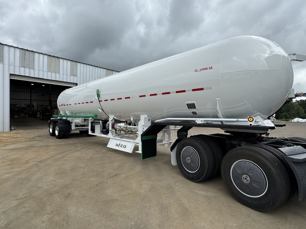 Large white tanker trailer with gauges and hoses parked outside a hangar under cloudy sky.