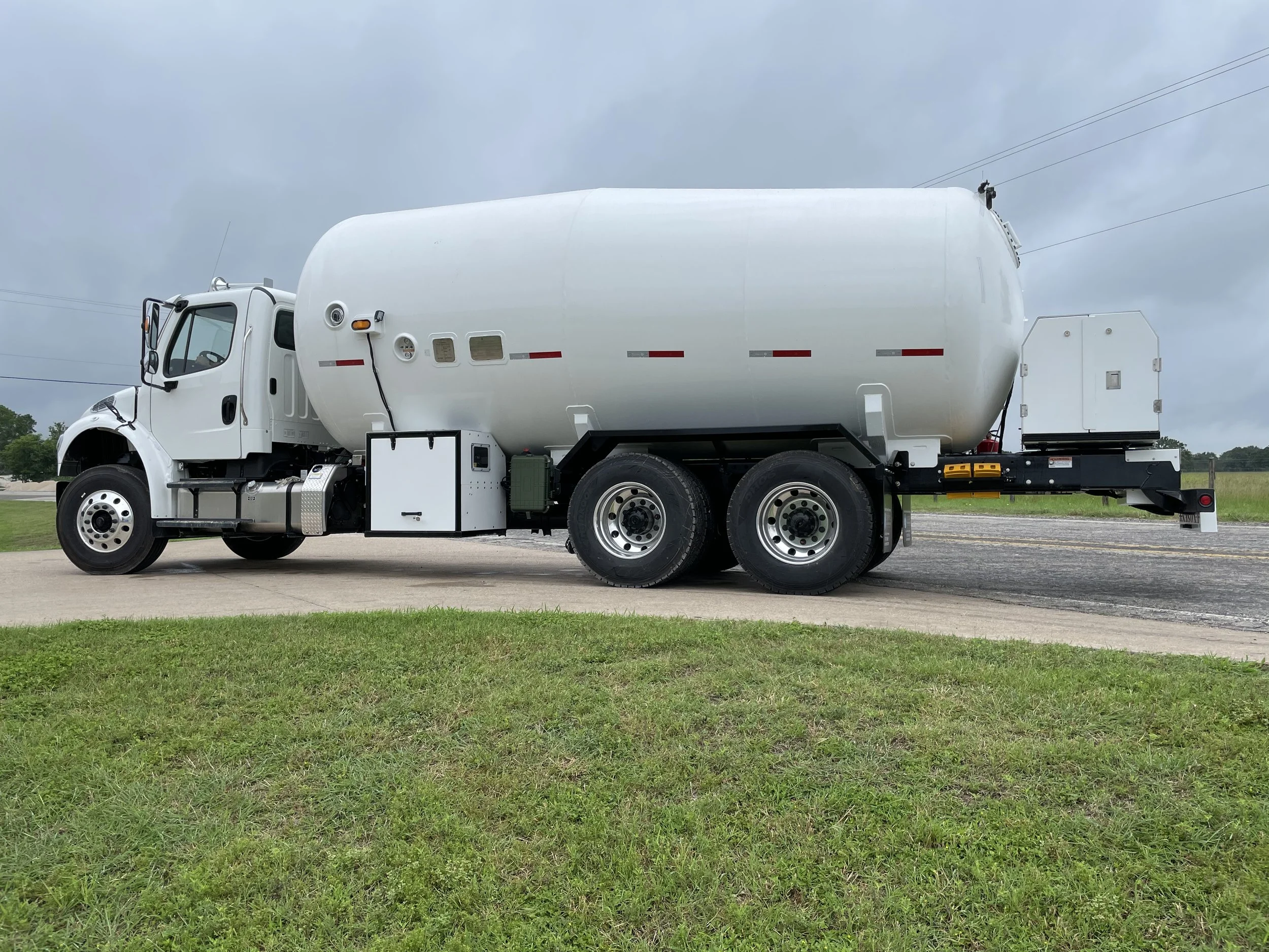 A large white tanker truck parked on a concrete driveway with green grass in the foreground and a cloudy sky in the background.