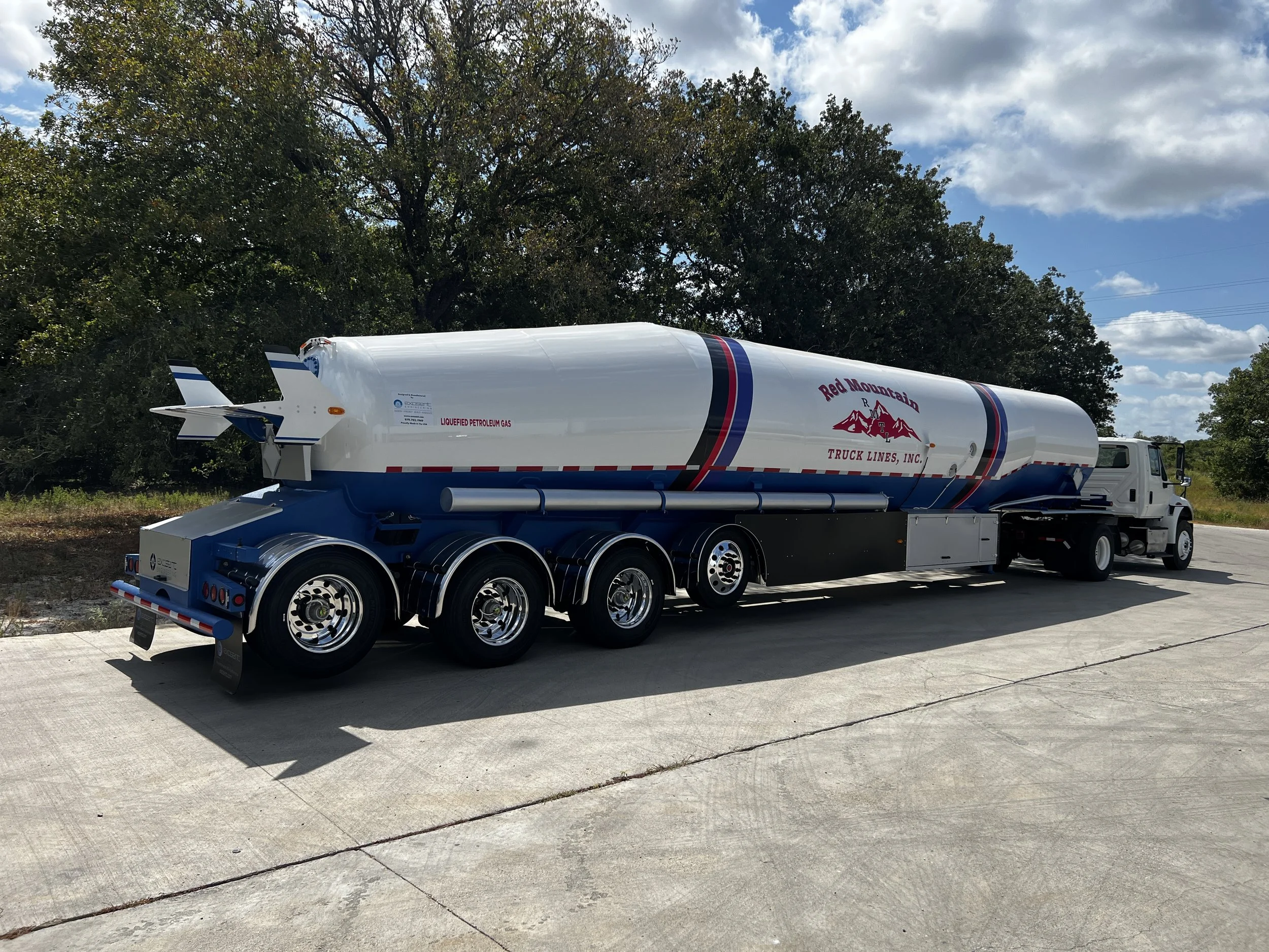 Fuel tanker truck with white, blue, and red branding for Red Mountain Truck Lines, Inc., parked on concrete pavement with trees in the background and a partly cloudy sky.