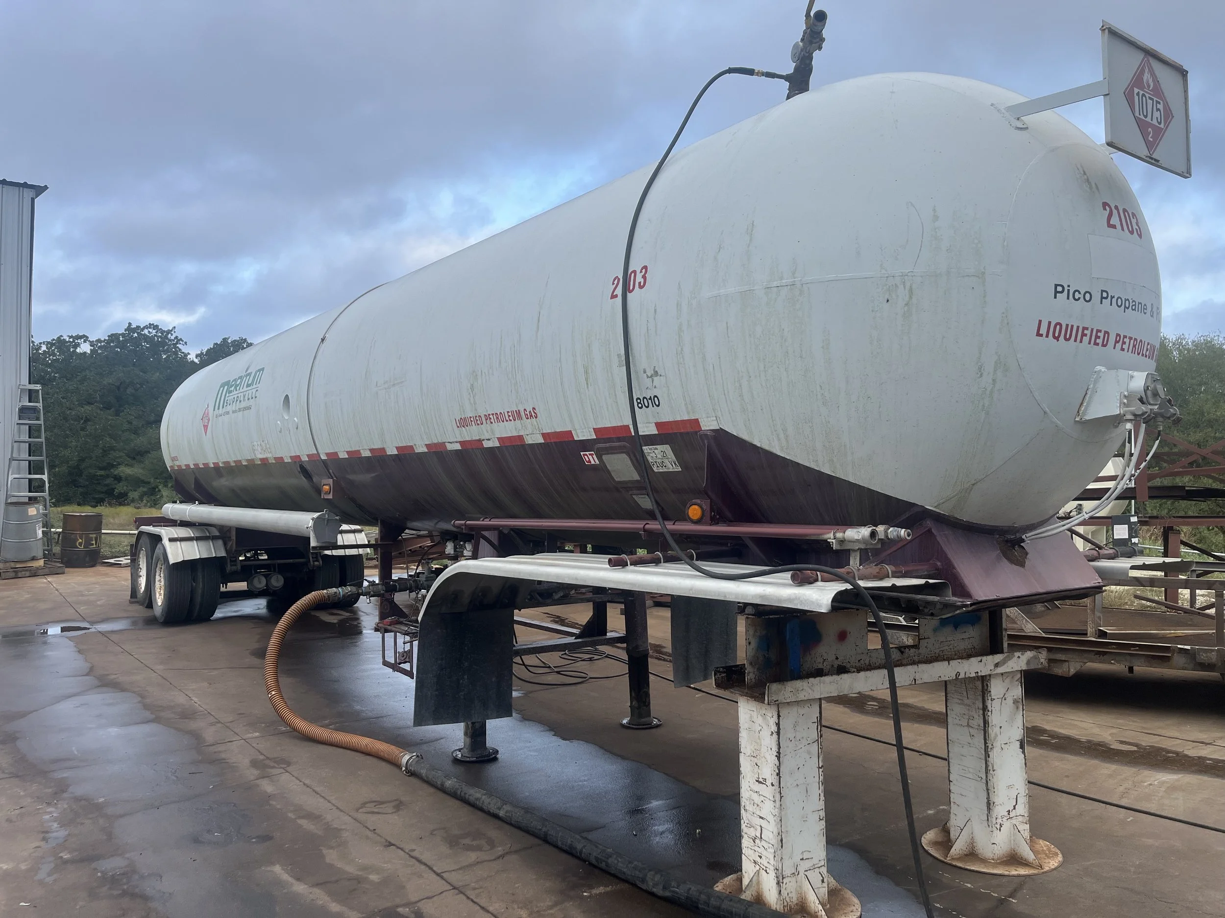 Large white liquefied petroleum gas tank on a platform outdoors, with hoses connected, against a cloudy sky and trees in the background.