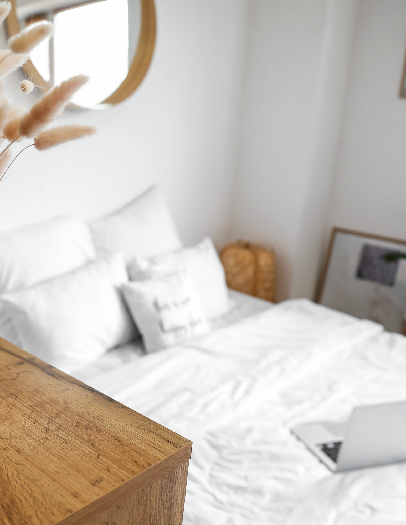 A cozy bedroom with a white bedspread, pillows, and a small open laptop on the bed. A wooden nightstand is next to the bed, and a wicker chair is in the corner. The wall decor includes a mirror and framed artwork.