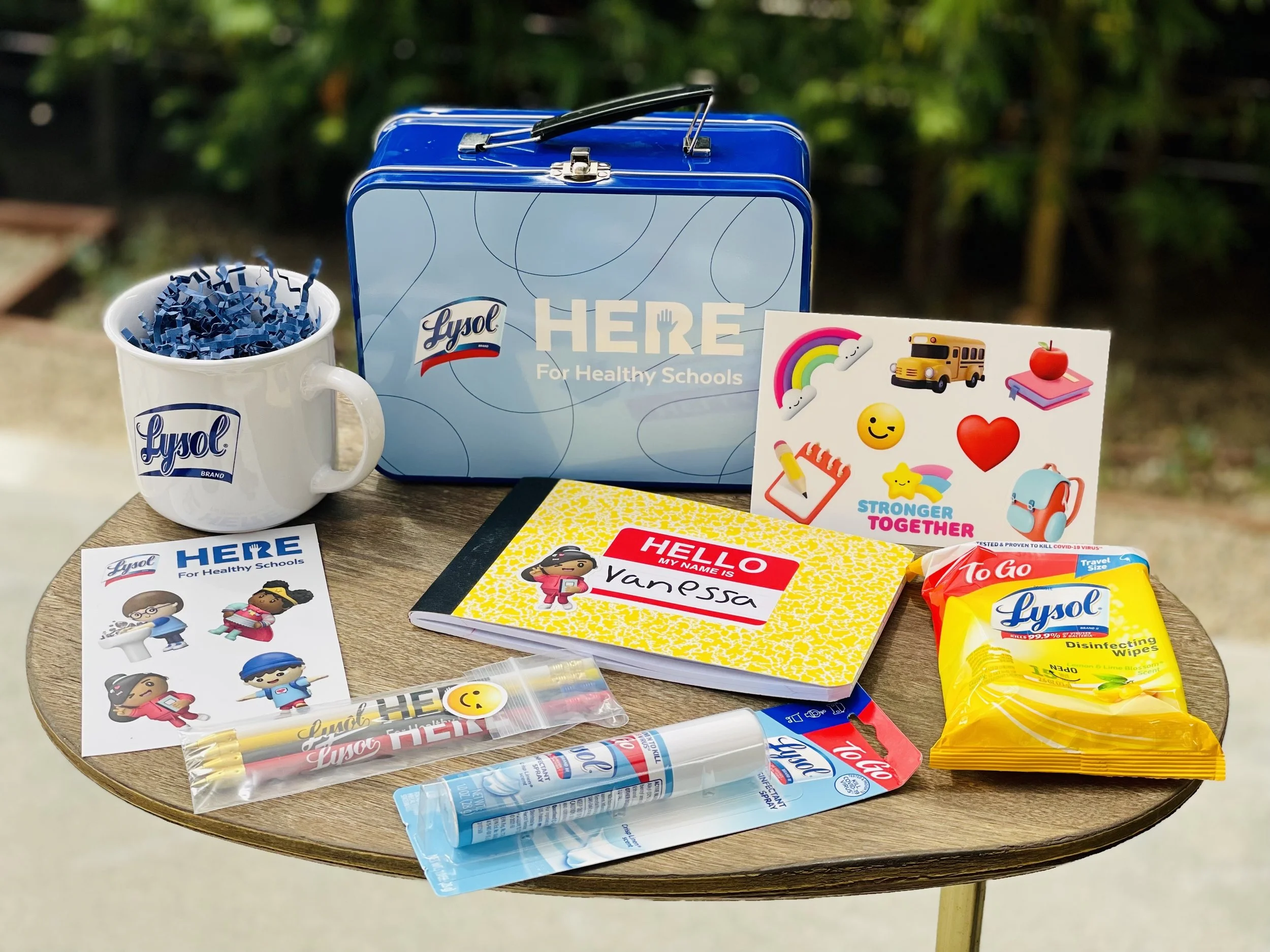 School supplies including Lysol disinfecting wipes, a mug with Lysol branding, a small metal tin, stickers with positive messages and images, a yellow pack of Lysol disinfectant wipes, a blue spray bottle, a yellow notebook, a sign that says 'HERE For Healthy Schools,' and other stationery on a wooden table outdoors.