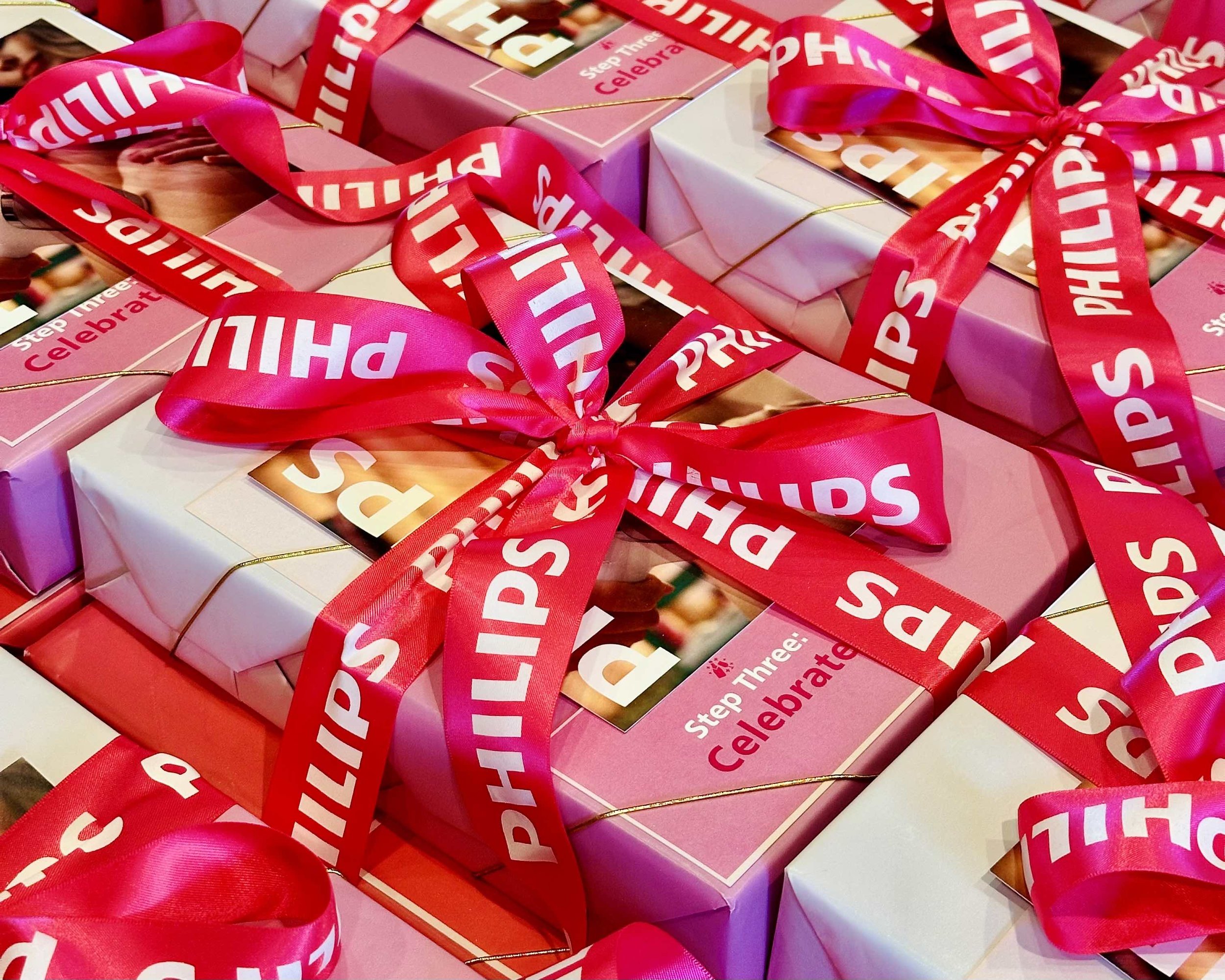 Close-up of wrapped gift boxes with pink, white, and purple wrapping paper, decorated with red ribbons and tags that say 'Step Three Celebrate'.