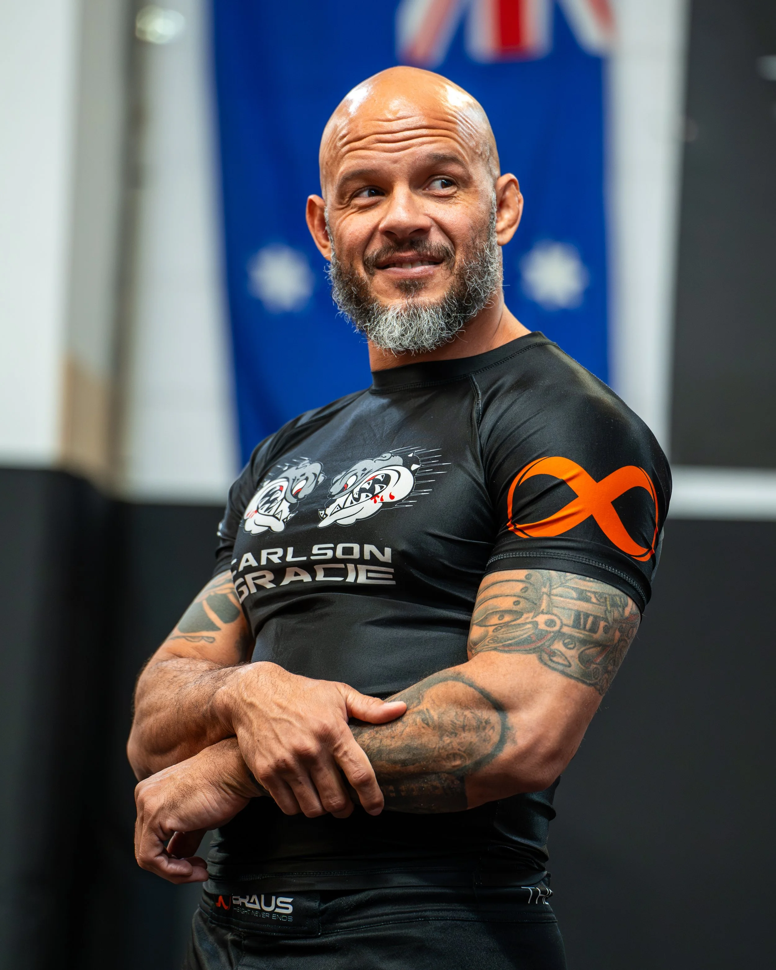 A bald man with a gray beard wearing a black athletic shirt with graphic designs and tattoos on his arm, standing in front of a blurred background with an American flag and blue backdrop.