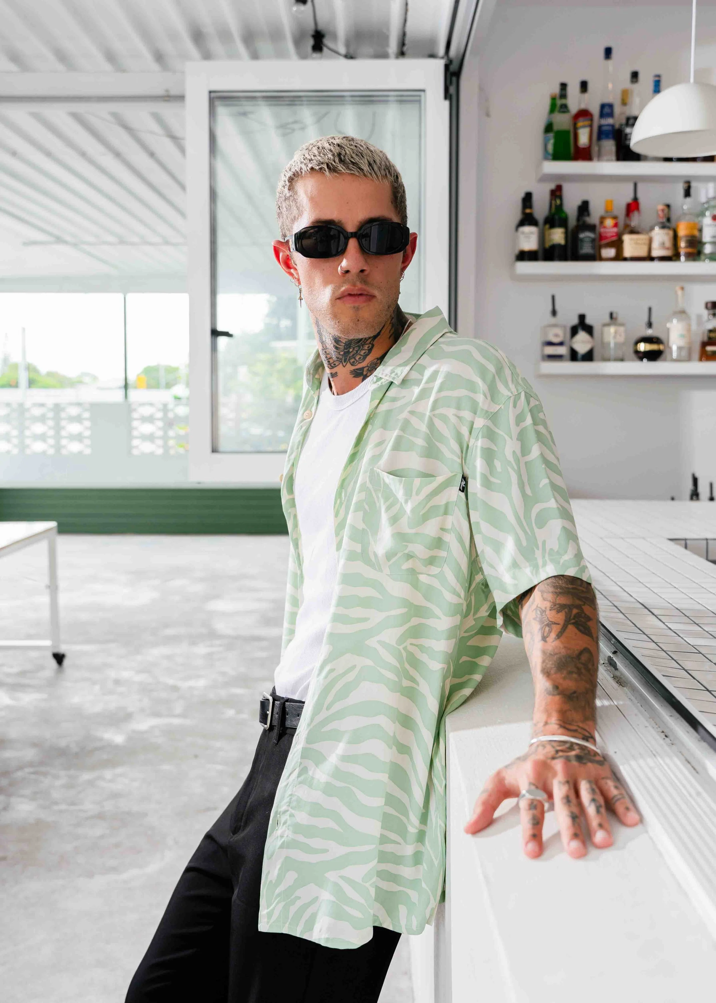 A young man with tattoos, wearing sunglasses and a light patterned shirt, is leaning on a white counter inside a modern kitchen or bar area.