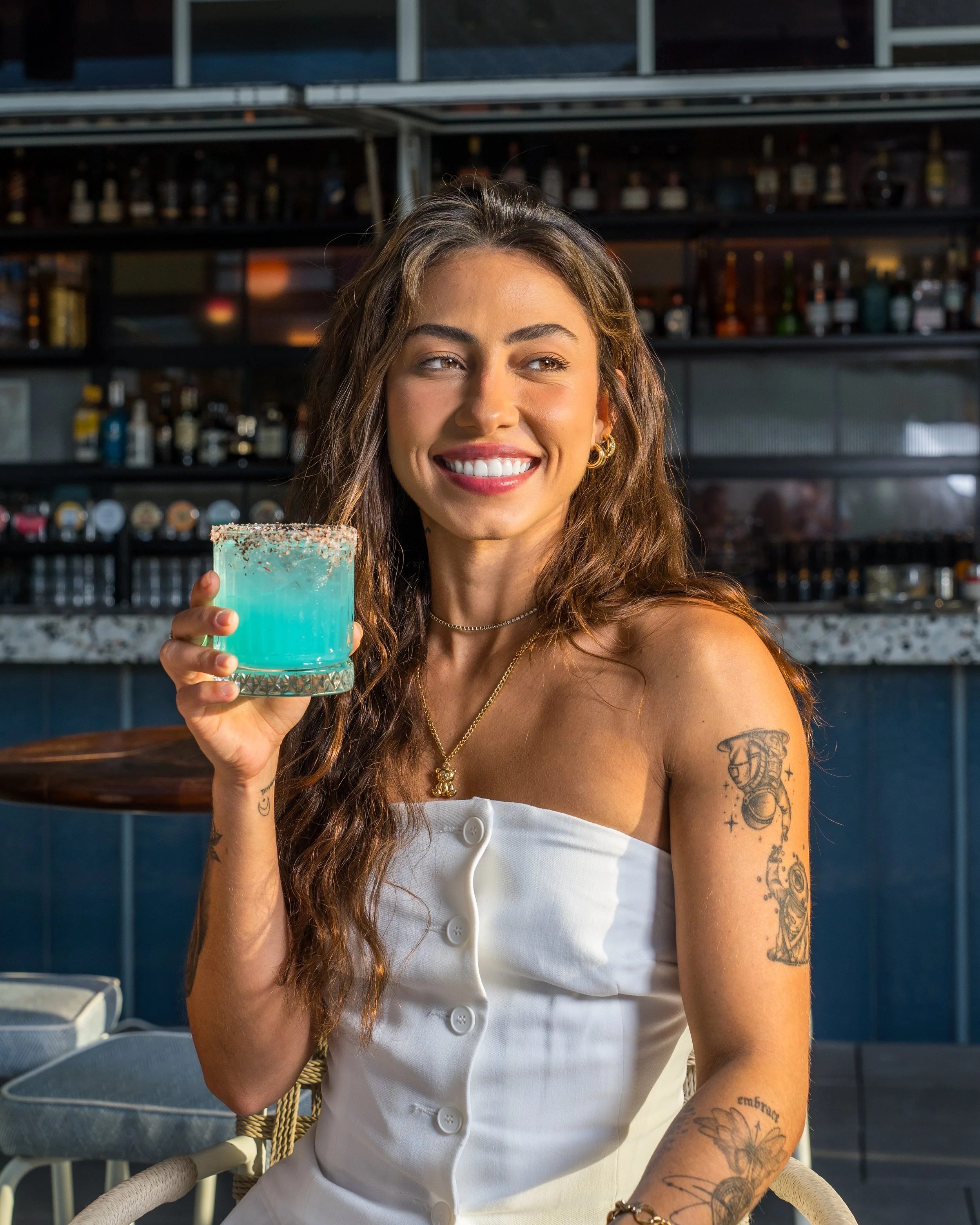 A woman with long wavy hair, tattoos on her right arm, and a necklace, is sitting at a bar or restaurant, holding a blue cocktail in a glass with sprinkles on top, and smiling.
