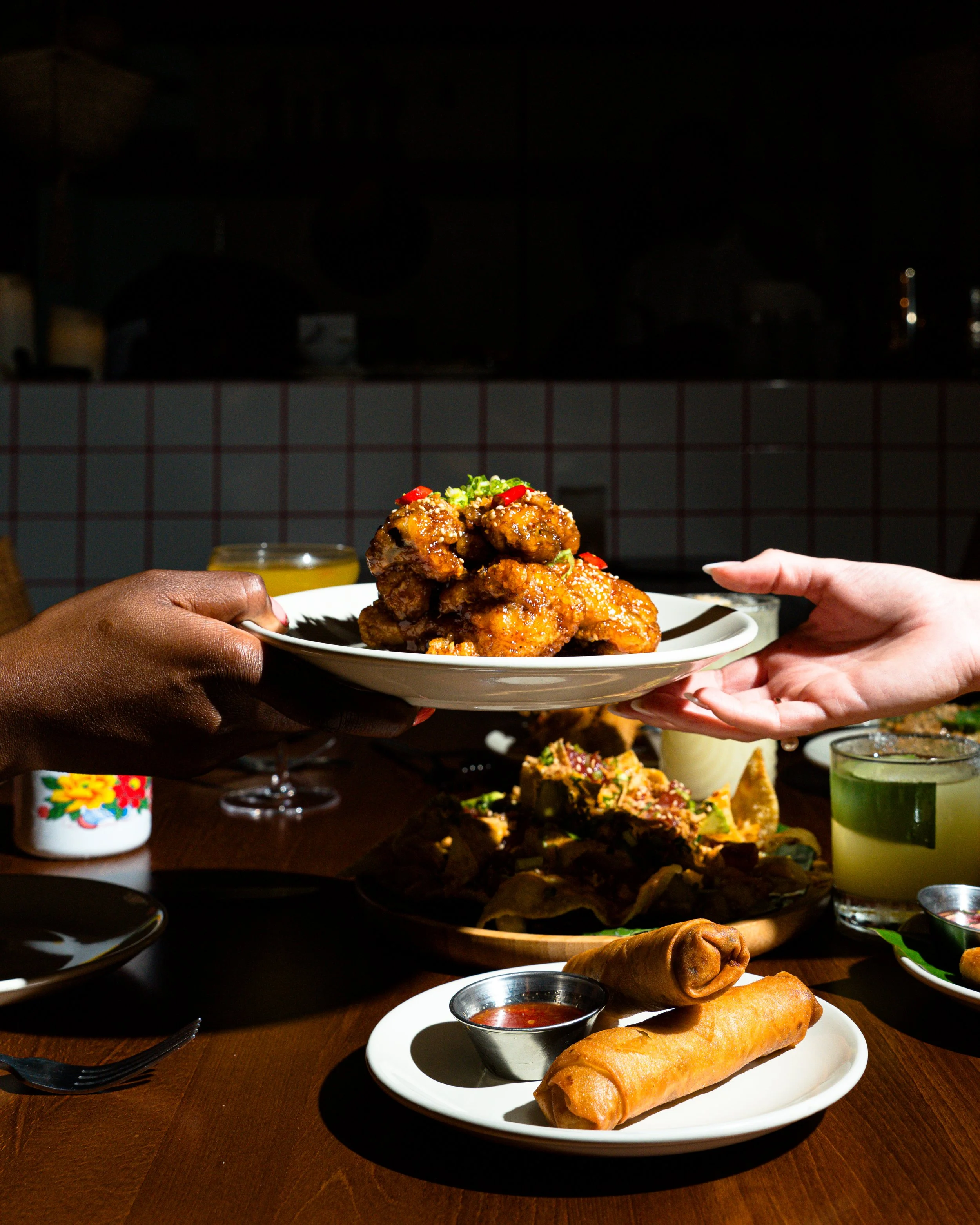 Two people exchanging a plate of fried chicken wings at a dinner table with various dishes and drinks.