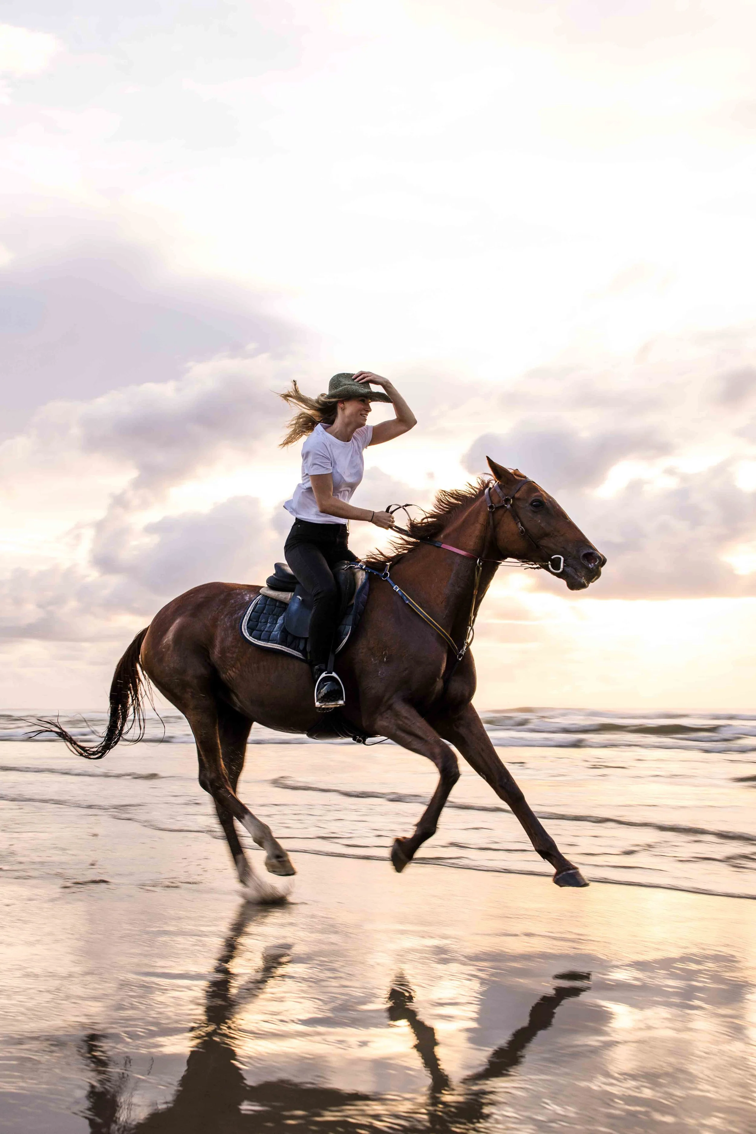 A young woman riding a brown horse on the beach during sunset, holding a cowboy hat in one hand and smiling.