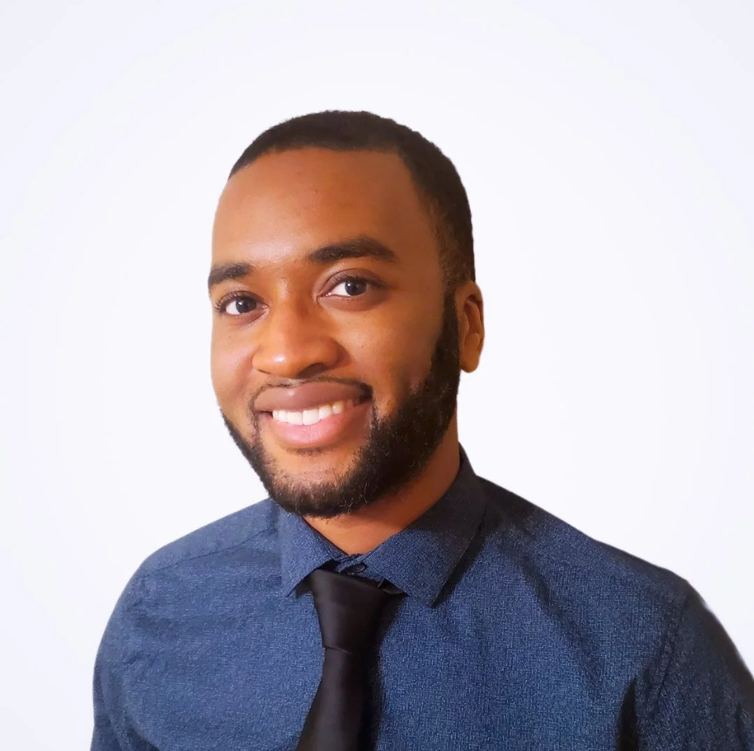 A smiling man with a beard, wearing a dark blue shirt and a black tie, against a plain white background.