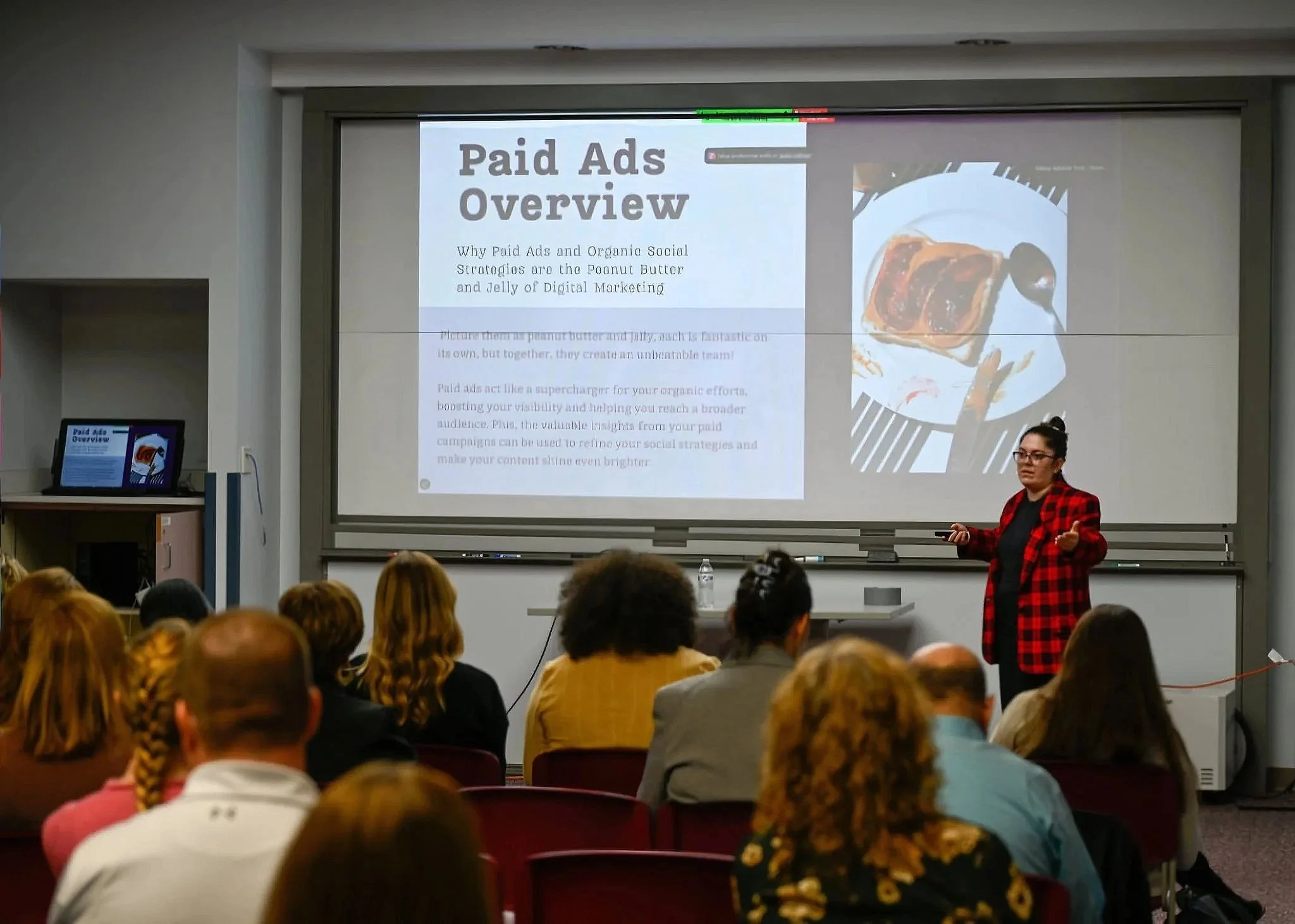 A woman in a red plaid blazer is giving a presentation in a classroom to an audience of adults. The presentation slide on the screen reads 'Paid Ads Overview' and discusses social media strategies. The room has a whiteboard and a laptop on a small desk.