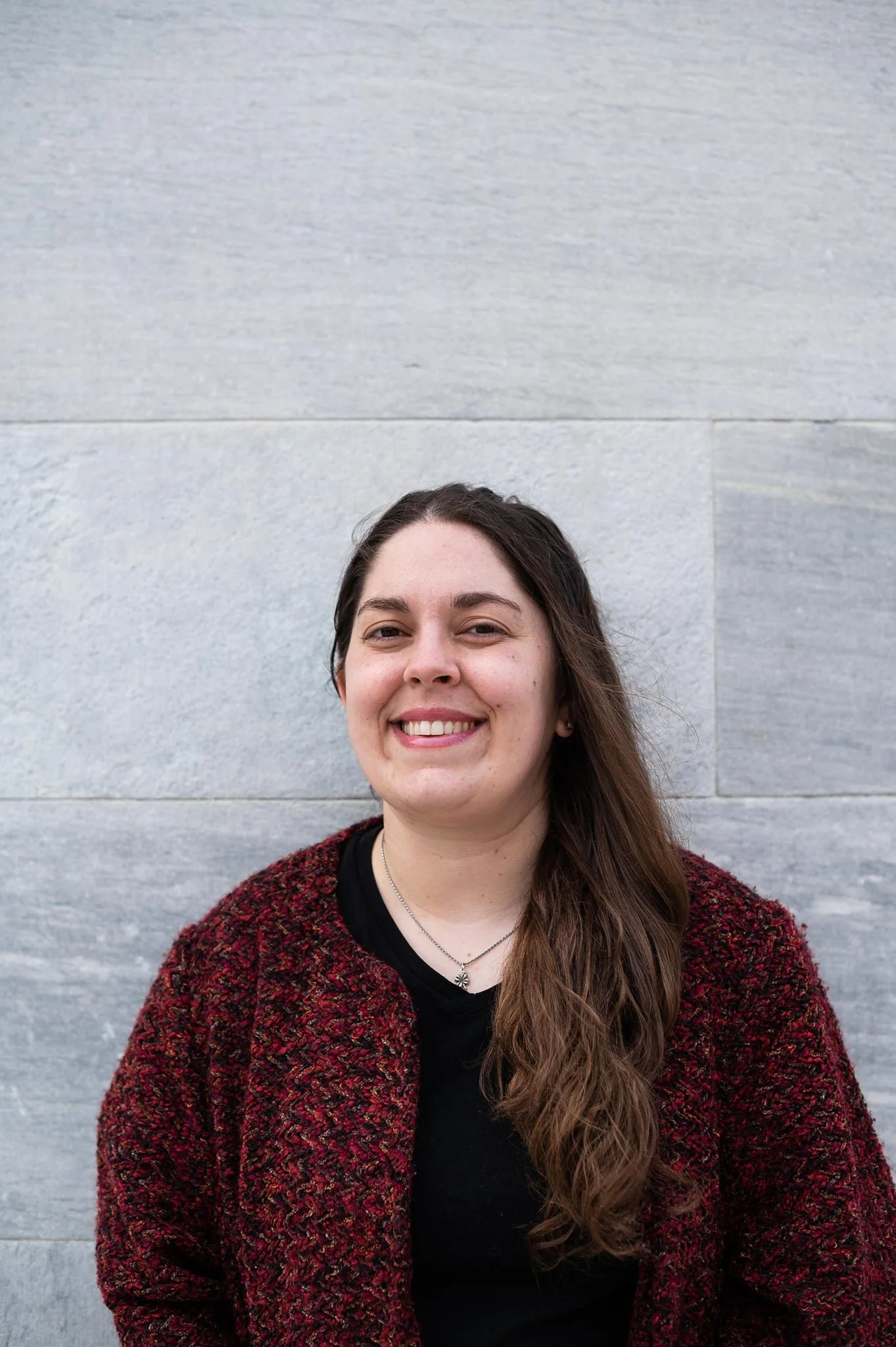 Sarah stands in front of a wall in a burgundy cardigan