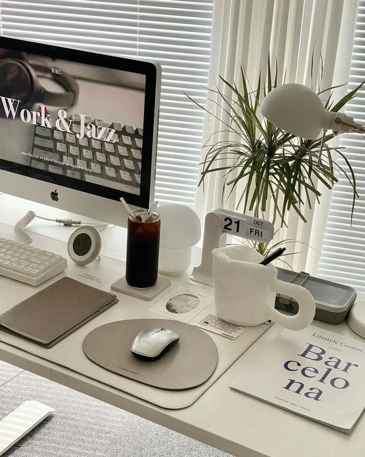 A white desk with an iMac computer displaying 'Work & Jazz', a plant with long narrow leaves, a calendar showing October 21, a glass of iced coffee, a cup of pens, a wireless mouse on a mouse pad, a closed notebook, a digital clock, a lamp, and a magazine titled 'Barcelon'.