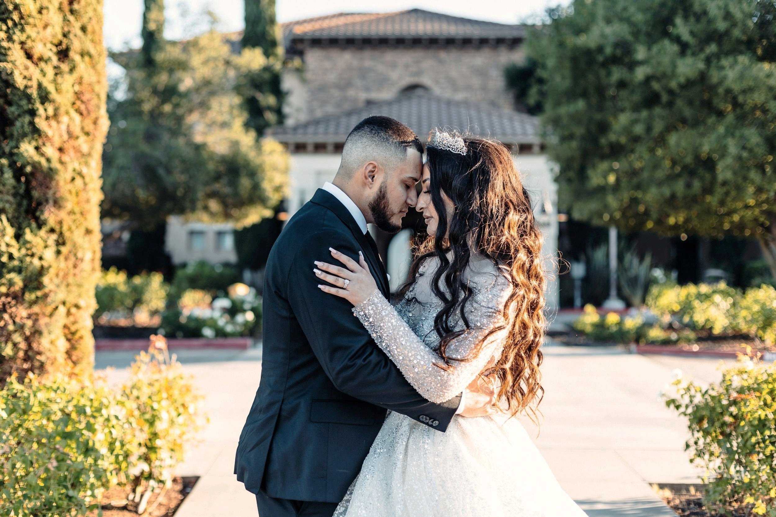 Bride and groom embracing outdoors, surrounded by greenery.