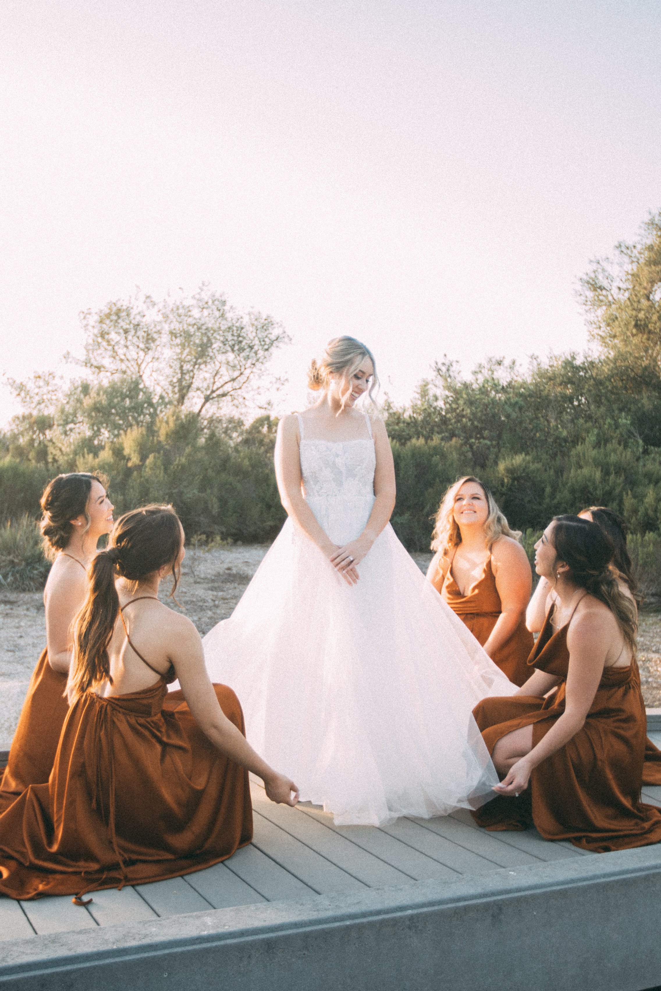 Bride in white gown surrounded by bridesmaids in brown dresses, outdoors.