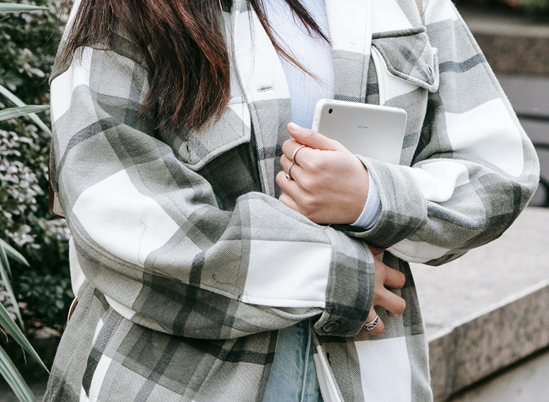 Person wearing a gray and white plaid jacket holding a smartphone outside near some greenery and a stone surface.