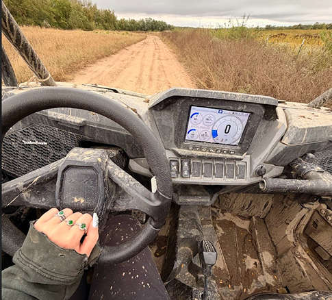 View from inside a muddy off-road vehicle on a dirt trail in countryside with fields and trees, showing the vehicle's steering wheel, dashboard, and part of a person’s hand on the steering wheel.