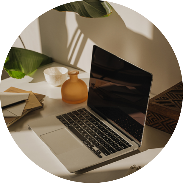 A laptop on a white desk surrounded by plants, a stack of papers, a notebook, and a small decorative vase with sunlight casting shadows on the wall.