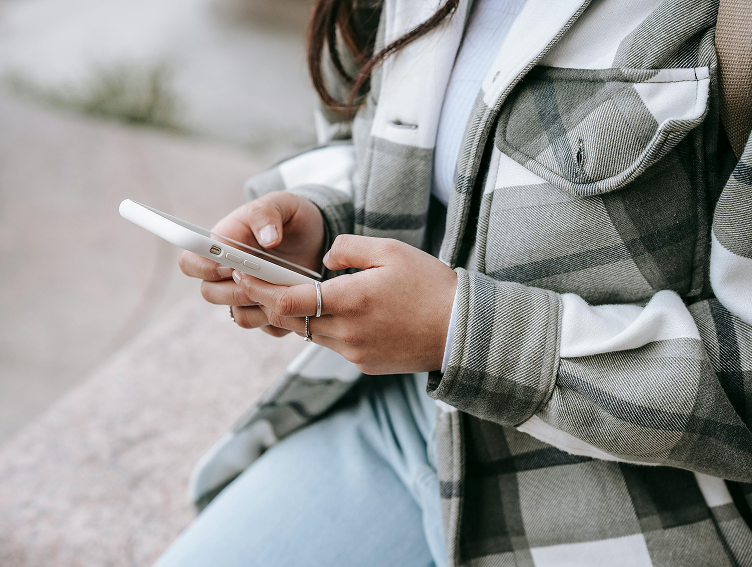Person in a plaid jacket holding a smartphone while sitting outdoors.
