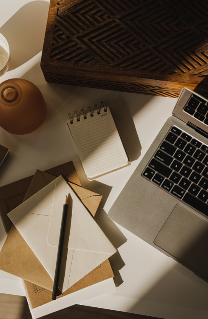 A cluttered workspace with a laptop, a small notepad, envelopes, a pen, a decorative vase, and a wooden box on a white desk.