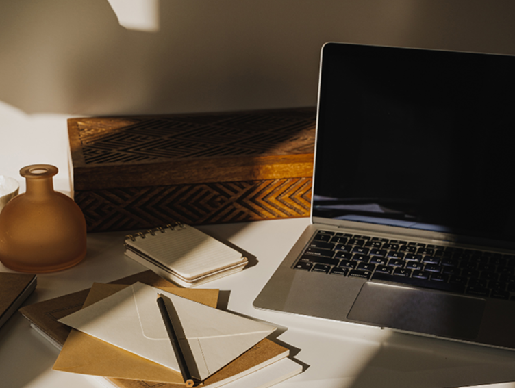 Laptop on a desk surrounded by notebooks, papers, a pen, a small notepad, and a decorative vase.