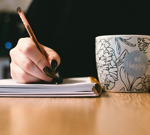 A person writing in a notebook with a pen, next to a ceramic mug that says "Be Happy" and has floral designs.