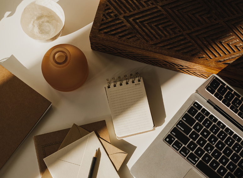 Desk setup with a laptop, notepad, envelopes, pen, two candles, a wooden box, and a notebook.