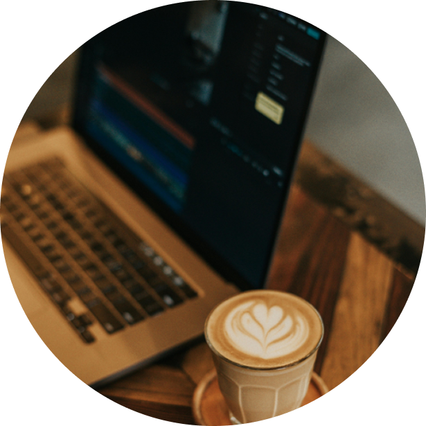 A laptop on a wooden desk with a screen displaying a dark-themed editing software, and a coffee drink with latte art in a glass mug on a wooden coaster in front of the laptop.