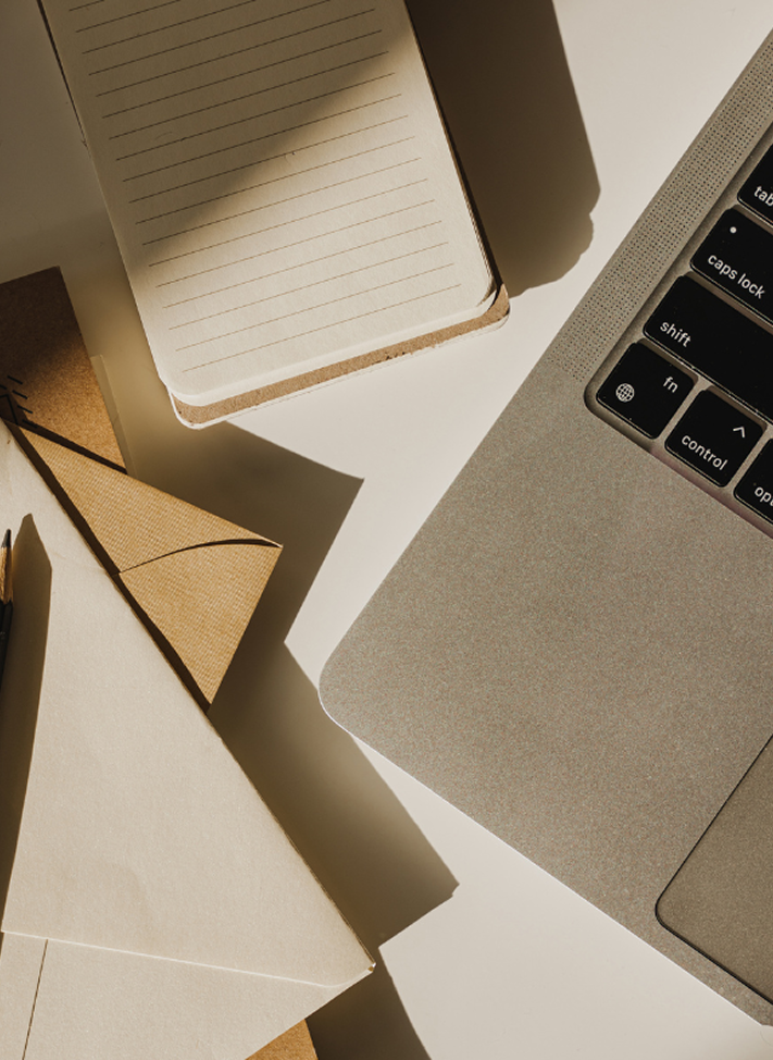 A workspace with a laptop, a small notebook, a pen, and some paper documents on a white surface, with sunlight casting shadows.