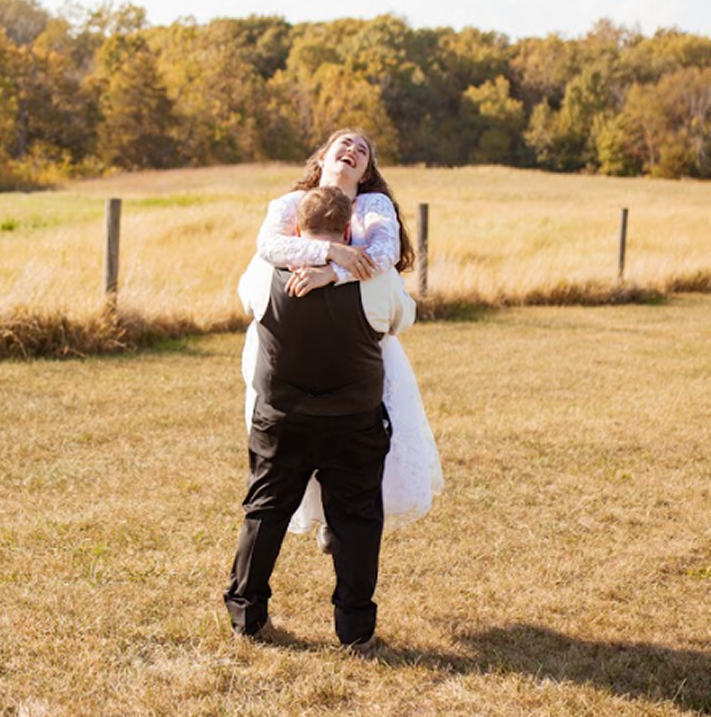 A couple enjoying a joyful moment outdoors in a grassy field. The woman, wearing a white dress, is being lifted by the man, who is dressed in black. The woman appears happy and is laughing, with a backdrop of trees and a sunny sky.