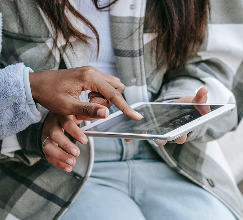 Two people, one with darker skin and the other with lighter skin, are using a smartphone together outdoors. The person with darker skin is holding the device, and the other is pointing at the screen.