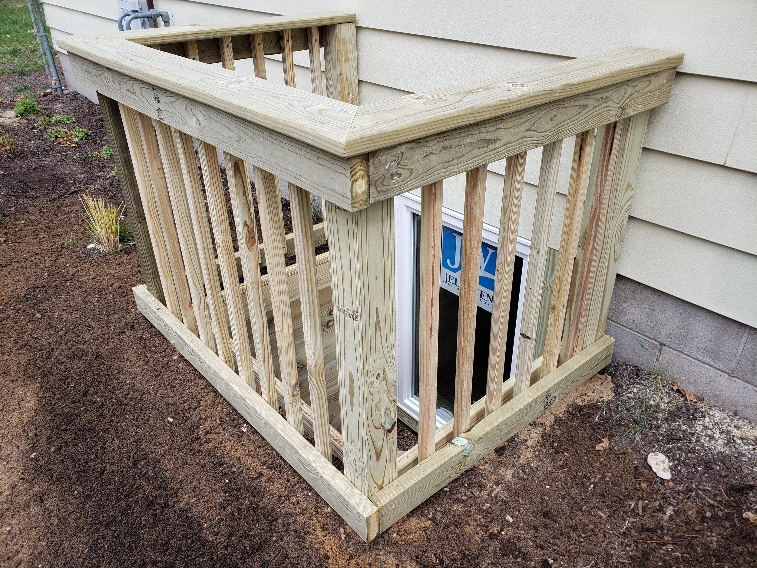 Unfinished wooden doghouse with a railing around the top, installed next to a house wall on dirt ground.