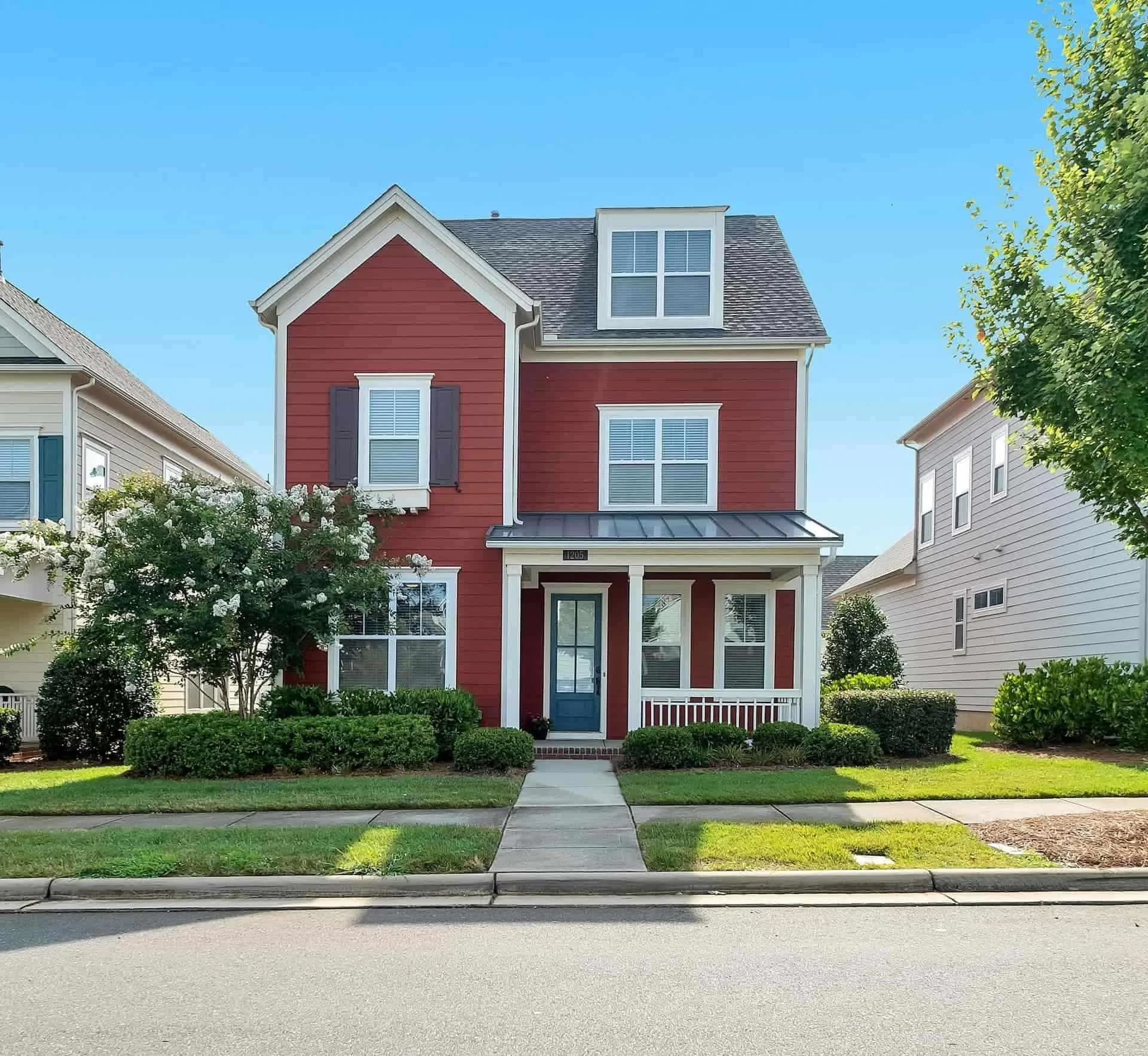 A red three-story house with a blue front door and white trim, surrounded by greenery under a clear blue sky.