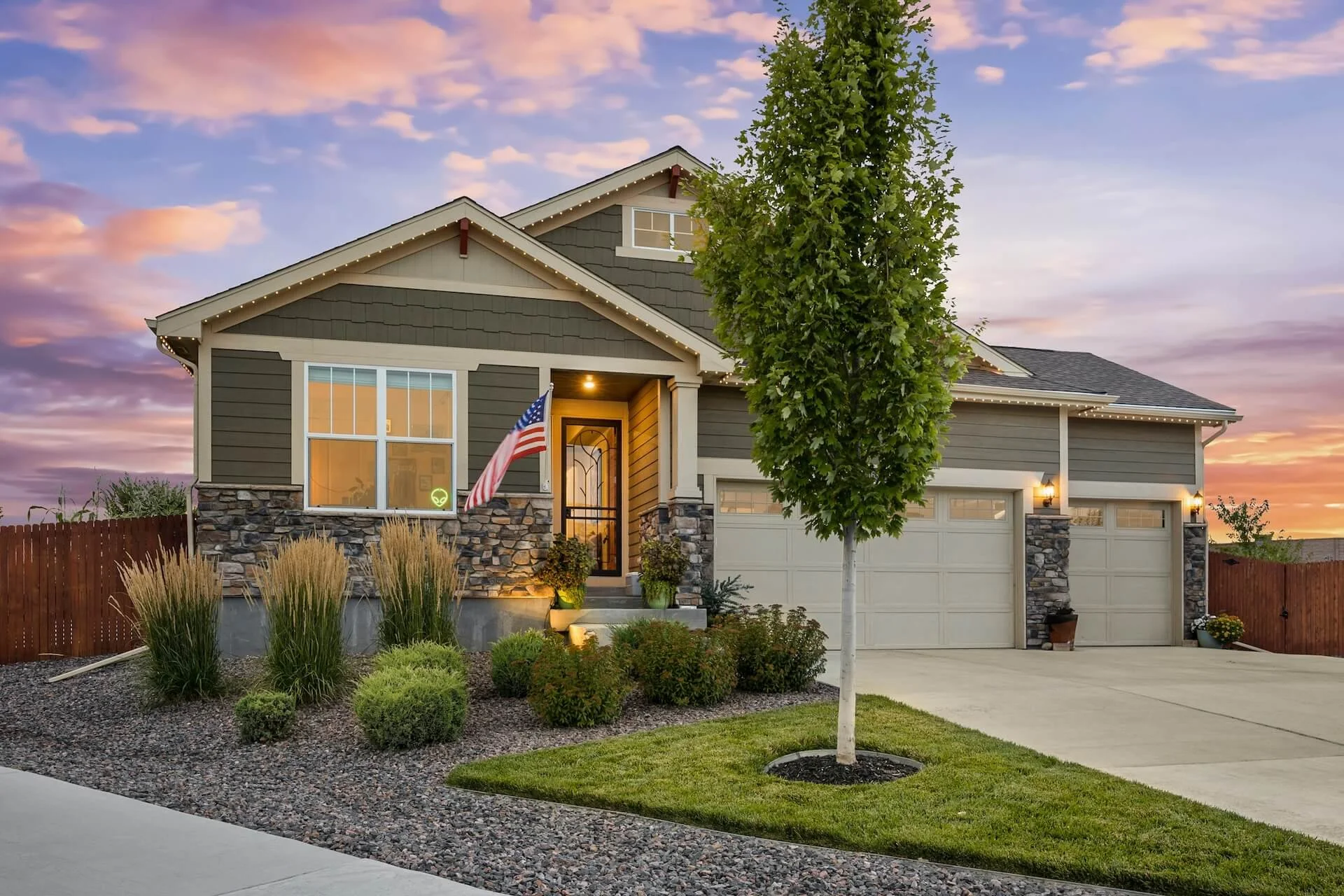 A modern, custom home build with a stone and siding exterior, American flag in front yard, surrounded by landscaped garden, trees, and flowers, during sunset.