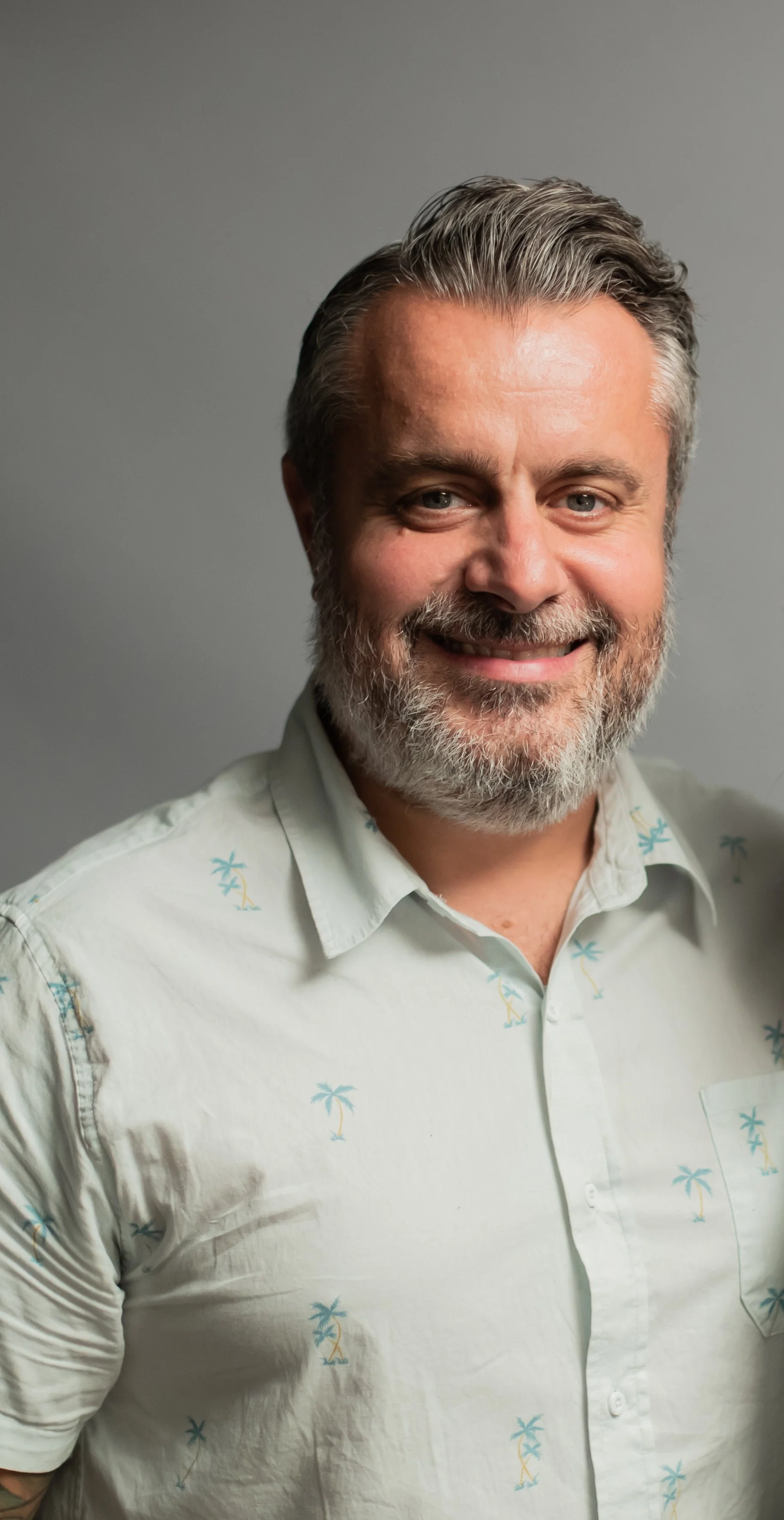 A smiling man with gray hair and a beard wearing a white short-sleeved shirt with blue palm tree patterns, standing against a plain gray background.