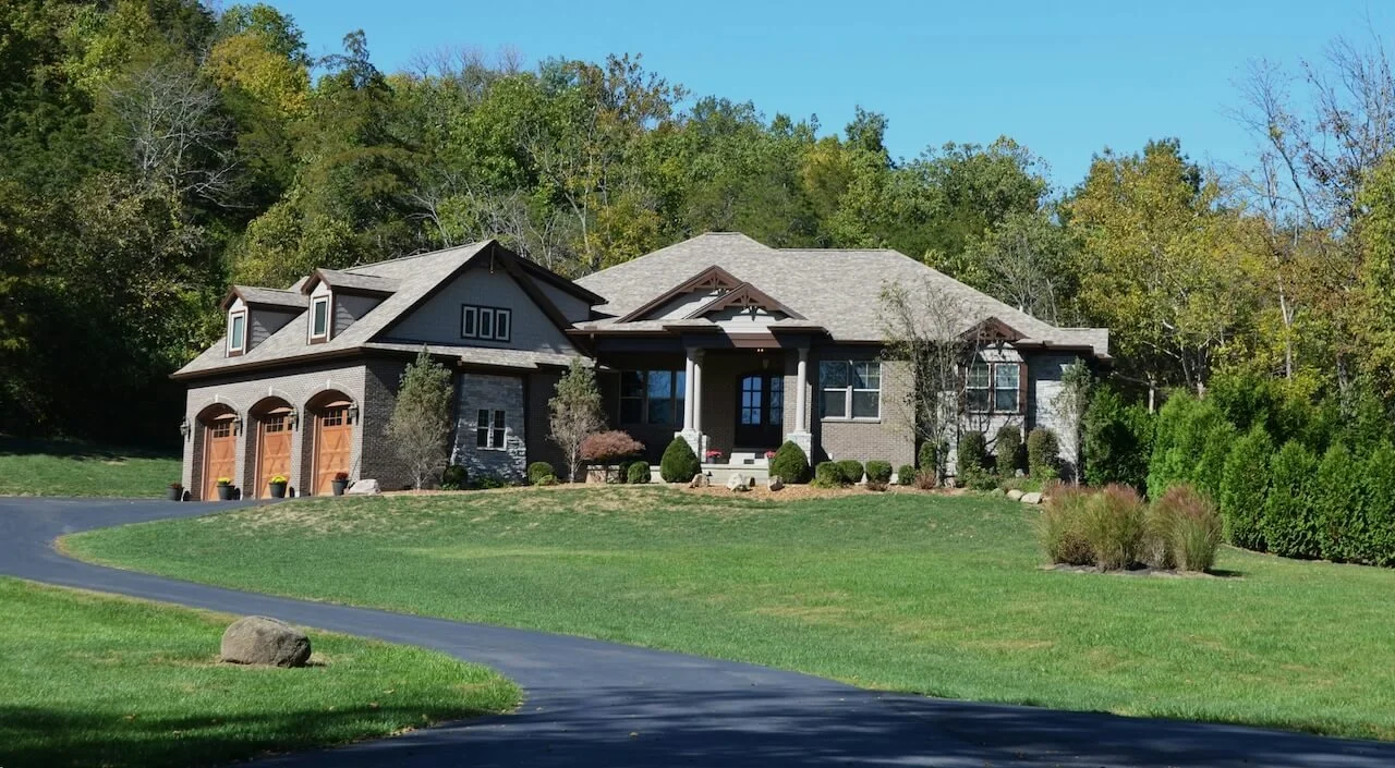 Large brick house with front porch, multiple windows, and attached three-car garage surrounded by green lawn and trees.