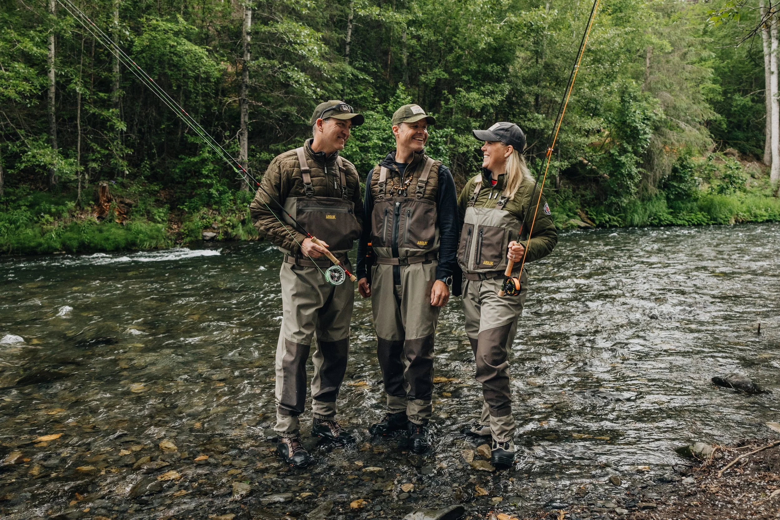 Three anglers standing in an Alaska river wearing Arolik waders, smiling and holding fly rods during a fishing trip.