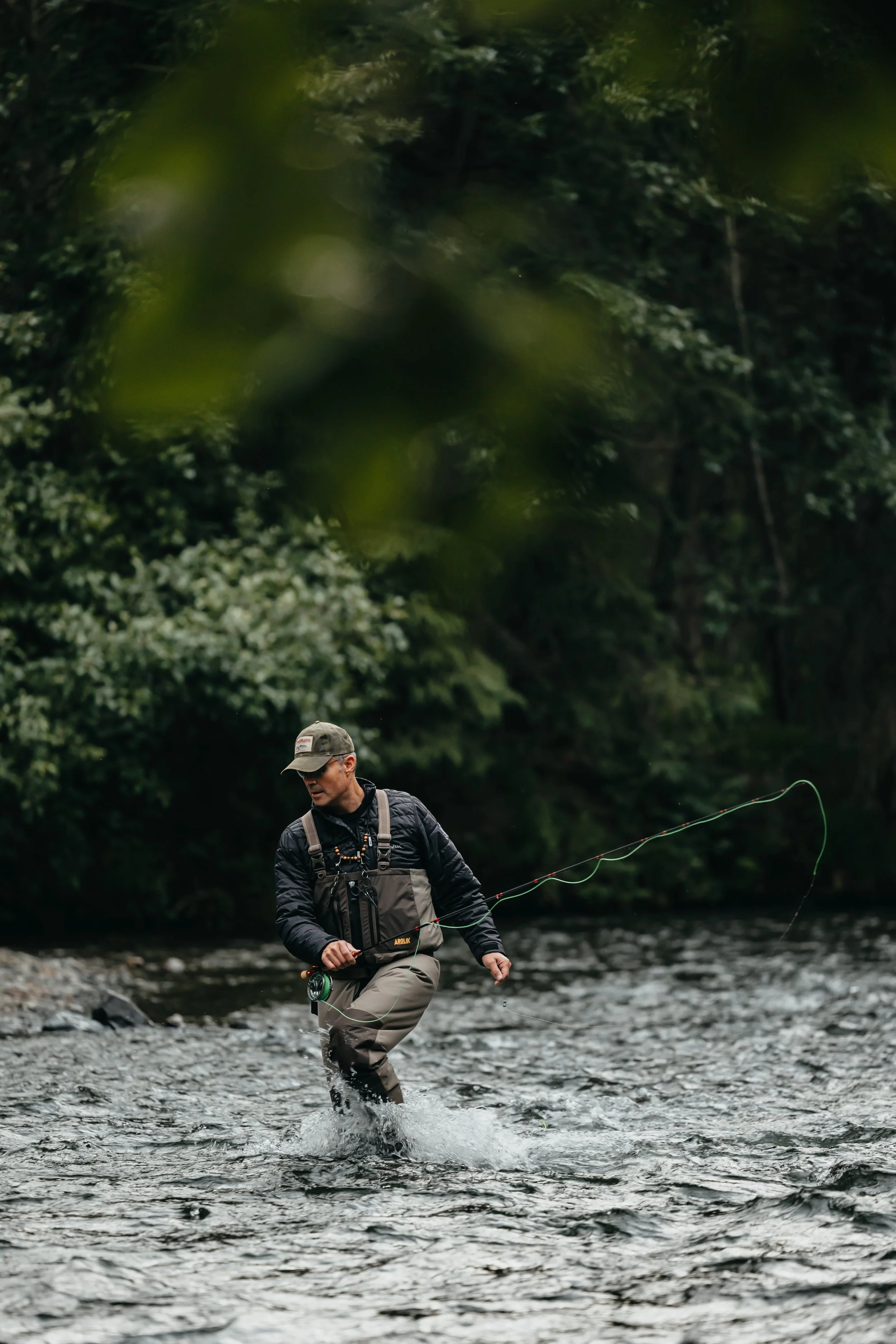 Man fly fishing in a river wearing Arolik waders, casting line in a moody forest setting in Alaska.
