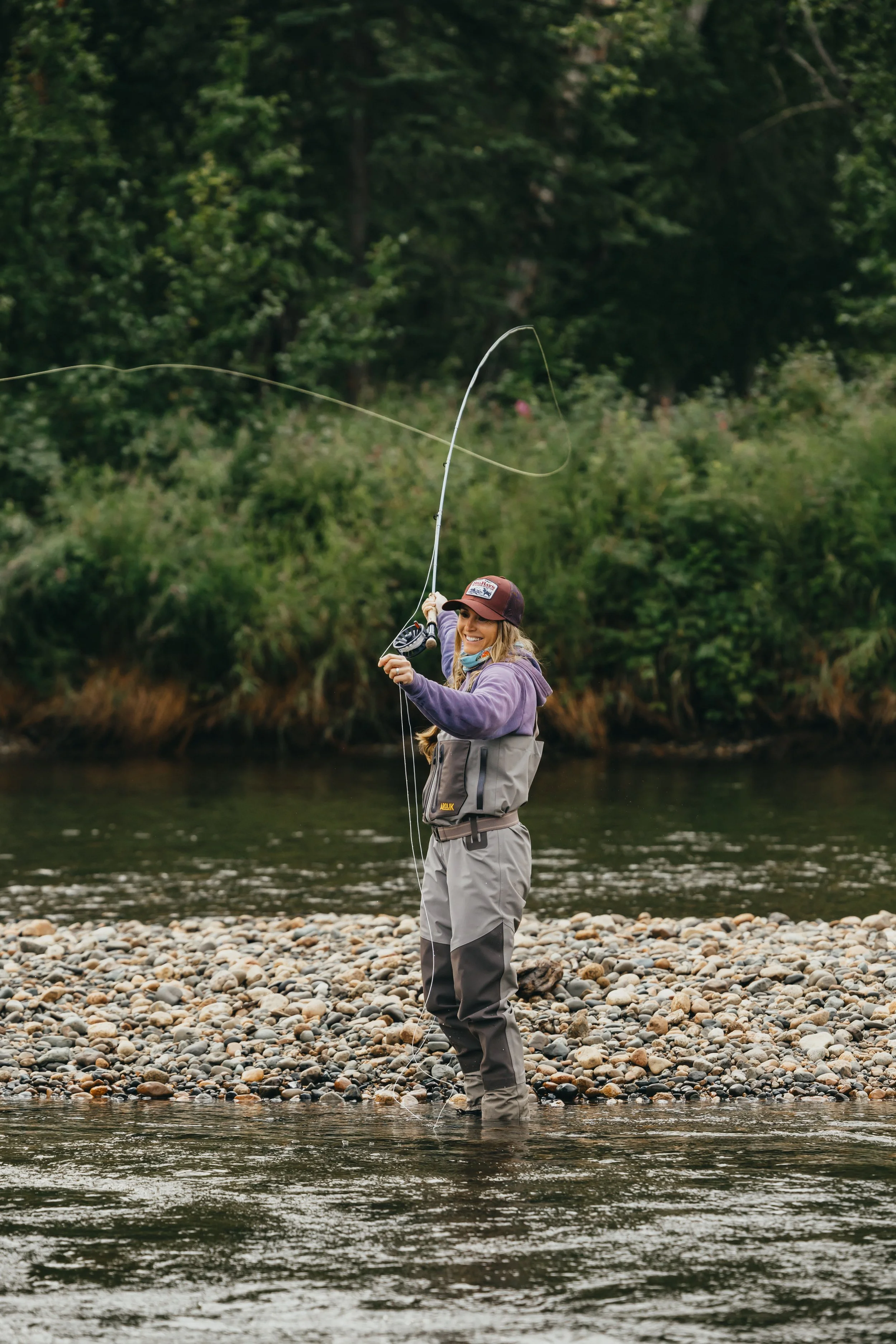 Woman fly fishing in Alaska river wearing Arolik waders, casting line in a natural wilderness setting.